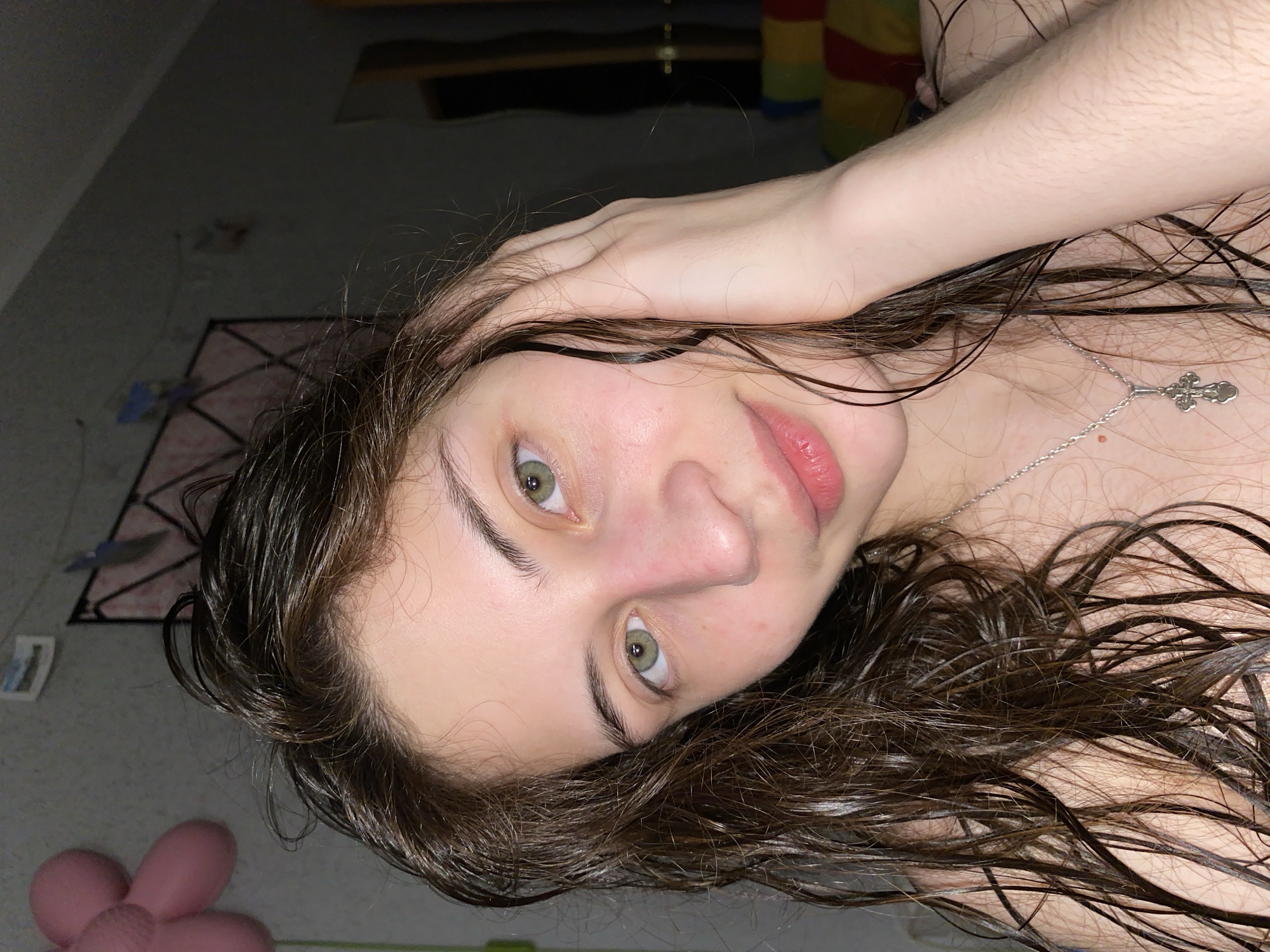 A young woman with wet, dark brown hair is lying down, looking up at the camera. She has light skin, green eyes, and is wearing a silver necklace with a cross pendant. Her hand is resting on her head.
In the background, a rug with a geometric pattern can be seen, along with a pair of pink slippers shaped like flowers. There is a closet with rainbow stripes visible above her hand.
The location cannot be verified as Votkinsk, Russia based on the visual data provided.