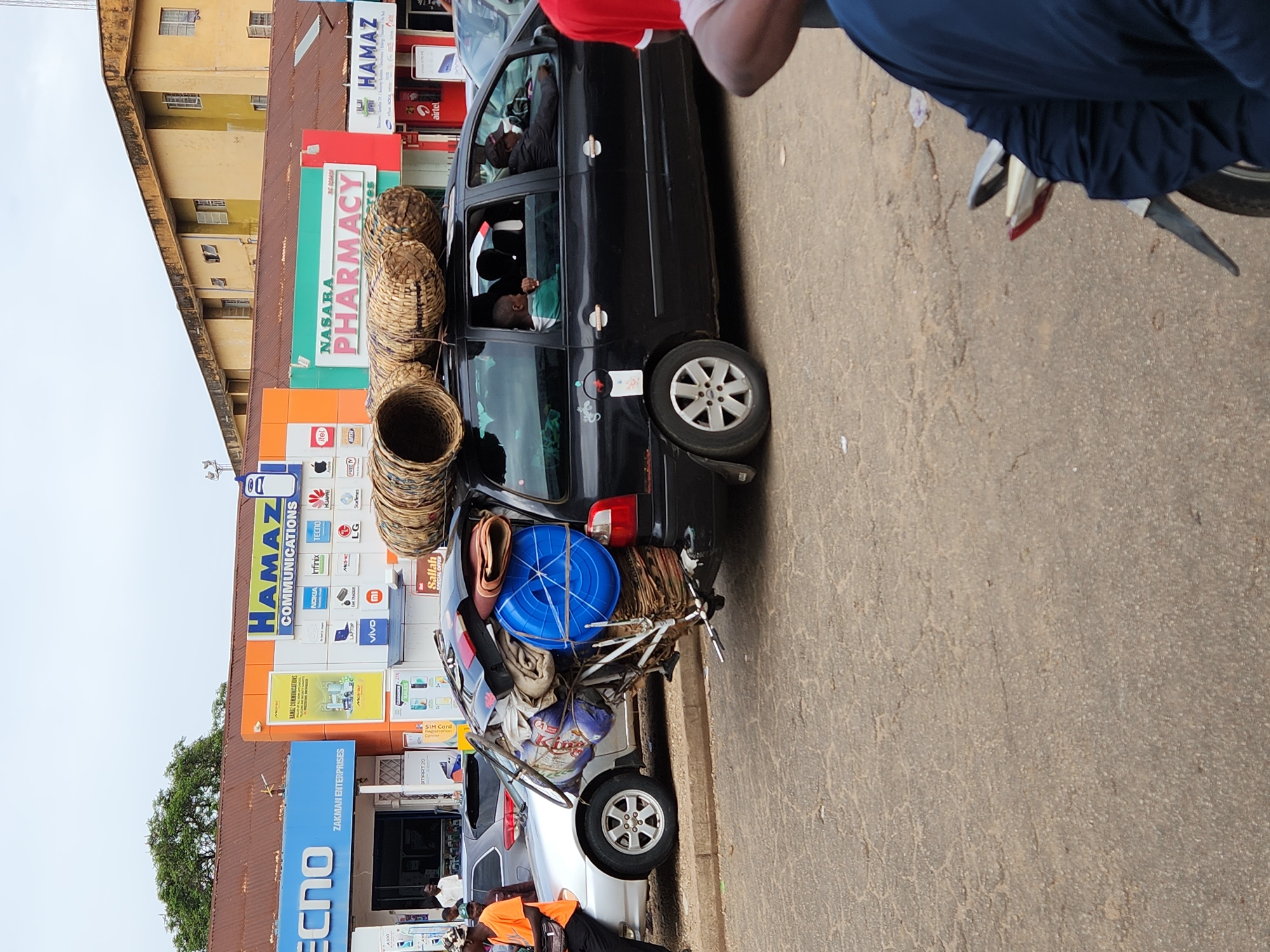 A street scene features a black car overloaded with goods, including baskets and a blue plastic container, strapped to its roof and rear. A silver car is parked behind it. The "NASARA PHARMACY" and "HAMAZ COMMUNICATIONS" signs are prominently displayed on the adjacent building. A person in an orange shirt stands near the silver car, while a person in a red shirt is partially visible near the black car. The street surface is gray and appears worn. The buildings have visible signage, and the general setting suggests an urban environment.