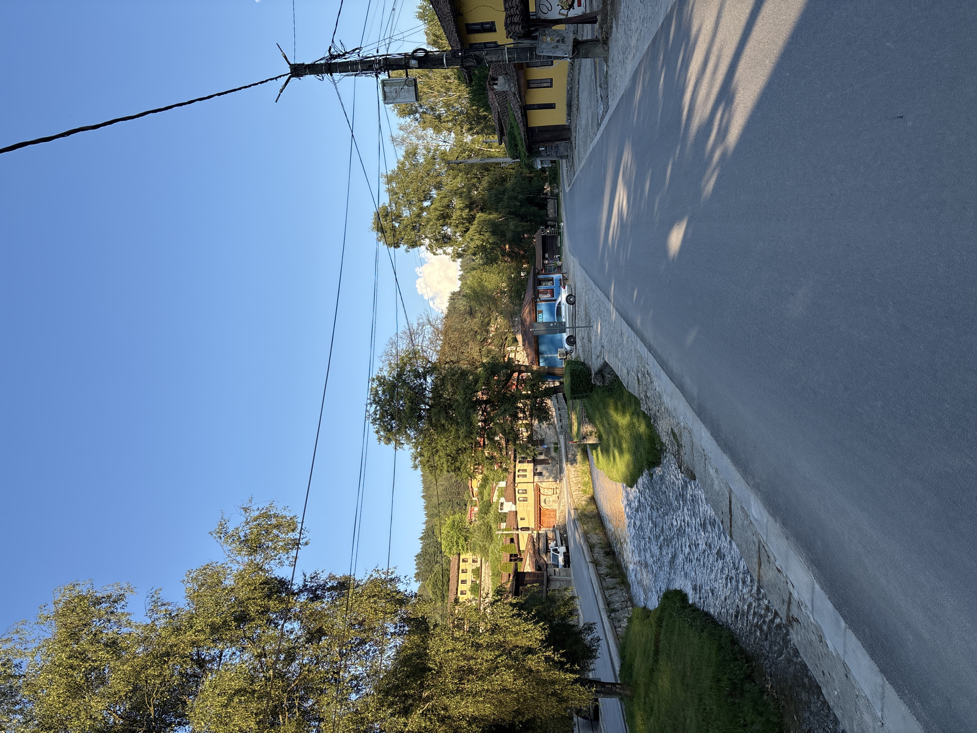 The image shows a street scene in Koprivshtitsa, Bulgaria. A paved road runs along the right side of the frame, bordered by a concrete embankment and a small, rocky stream. On the left side of the road, a village with traditional yellow houses and red tile roofs is visible, nestled against a tree-covered hillside. A dense canopy of green trees dominates the left side of the image, with some of the trees extending over the road. Overhead, a complex network of utility wires crisscrosses the bright blue sky, supported by wooden poles. The sunlight casts long shadows from the trees onto the road, suggesting it is late afternoon or early morning. The overall atmosphere is peaceful and serene, capturing a glimpse of daily life in a historic Bulgarian town. There is no visible text in the image.