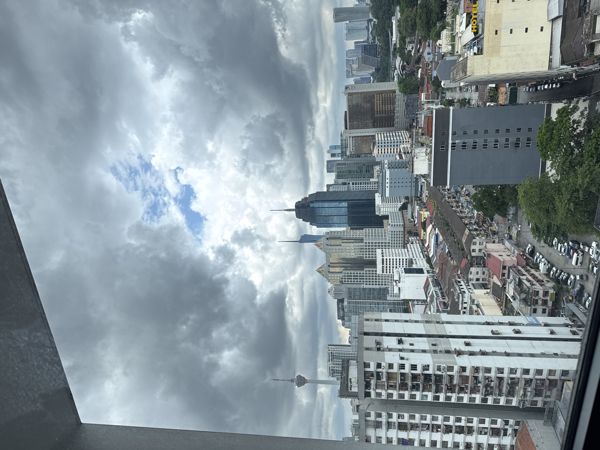 This is a high-angle, portrait-oriented, full shot of a cityscape under a cloudy sky. The scene depicts a dense urban environment with numerous tall buildings of varying architectural styles and heights. In the mid-ground, a distinct tower with a rounded top is visible, hinting at a communication tower or landmark. The buildings in the foreground appear to be residential apartments with many windows and balconies, some showing signs of air conditioning units and what appear to be personal belongings. Further back, modern skyscrapers with glass facades dominate the skyline, interspersed with older, lower-rise buildings. A street with parked cars is visible between some of the buildings.
The weather appears overcast, with heavy grey clouds dominating the sky, though patches of blue sky and lighter clouds are also present, suggesting a dynamic weather system or a break in the clouds. The lighting is diffused, typical of an overcast day. There is no clear indication of a specific time of day, but the overall scene suggests it is daytime. The image's composition, with the strong vertical lines of the buildings and the dramatic sky, evokes a sense of the scale and density of a major metropolitan area.
Visible text includes "HOTEL" in yellow lettering on a sign of one of the buildings in the mid-ground. The location is identified as Kuala Lumpur, Malaysia. The scene itself is a general view of urban life, with no specific activity or event prominently displayed. The focus is on the built environment and the sky.