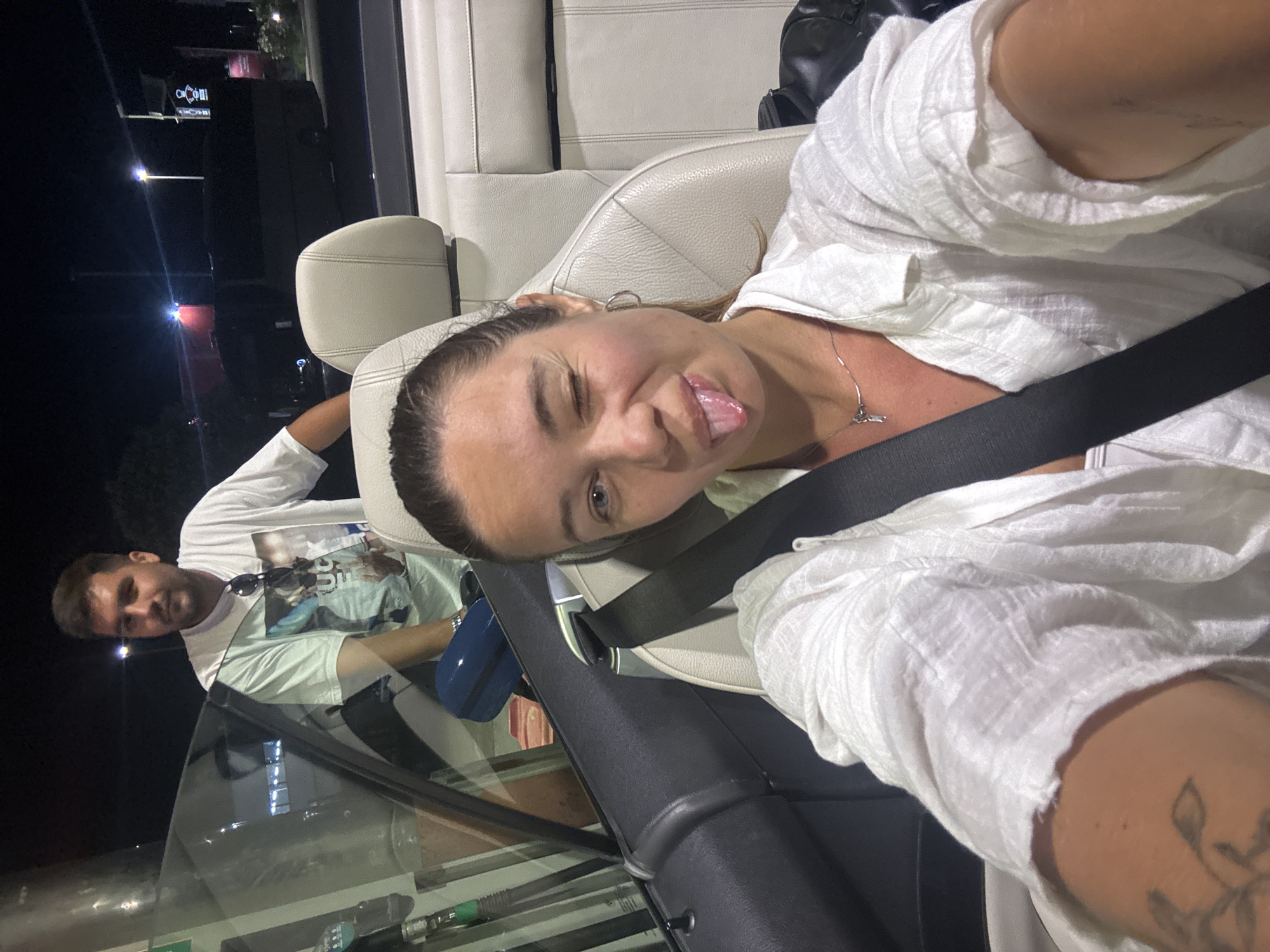 The image captures a young woman in the driver's seat of a convertible car, winking and sticking her tongue out at the camera. She is wearing a white linen shirt and a necklace. A man is visible in the passenger seat behind her, looking out the window and wearing a white t-shirt. The car appears to be at a gas station, with a gas pump visible to the left of the frame. The scene is at night, with streetlights and distant buildings illuminated. The location is identified as a gas station in Dub, Montenegro.