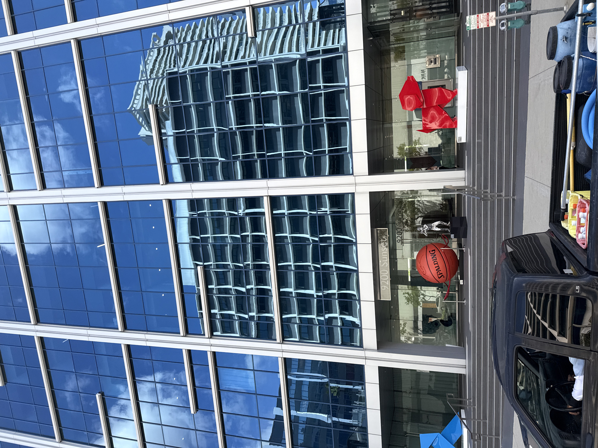 The image captures the entrance of the building at 9200 Sunset Boulevard in West Hollywood, USA. A modern glass-fronted building reflects an adjacent structure in its windows. The building's entrance features a large Spalding basketball on a stand, and a red origami-style sculpture above the doorway. A dark-colored vehicle is parked at the curb, with items such as blue hoses and cleaning supplies visible in the truck bed. The environment suggests a sunny day in a commercial urban area.