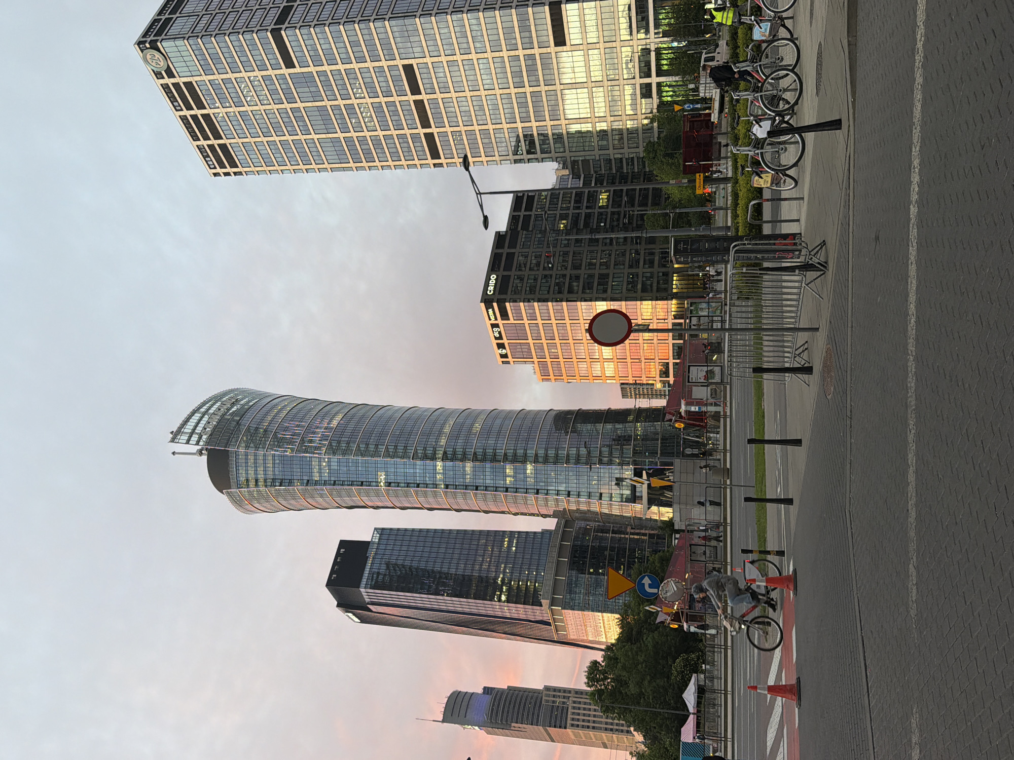 The image depicts a cityscape, likely captured at dusk or dawn given the soft pink and orange hues in the sky. Tall, modern skyscrapers with glass facades dominate the scene. One building has a distinctive curved shape, making it stand out from the others. Street level includes a paved walkway or road, with bicycles parked along the side. There is also some road signage and traffic cones. The overall impression is one of an urban setting with a mix of commercial and public spaces.