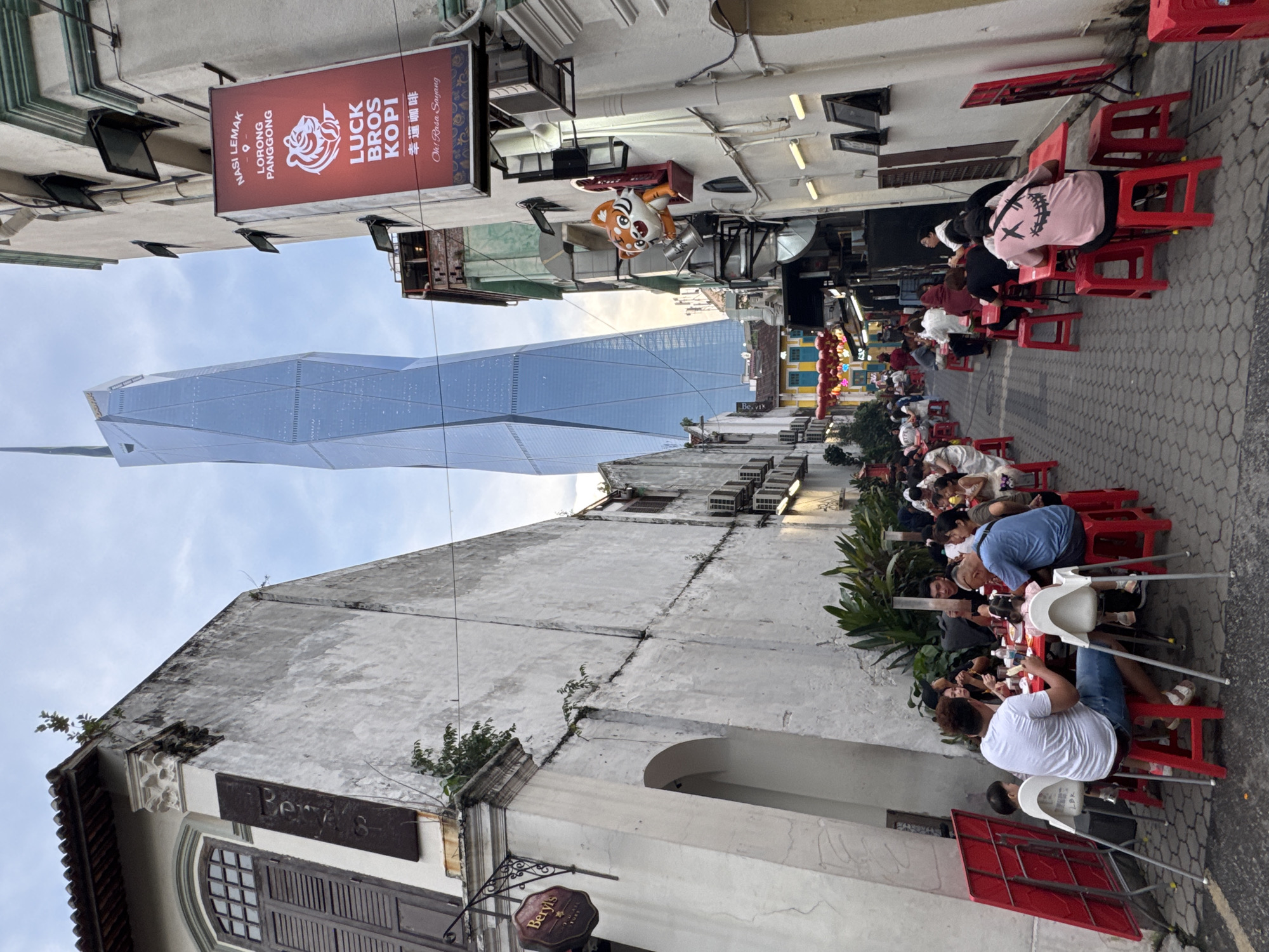 The image captures a narrow street scene in Kuala Lumpur, Malaysia, juxtaposing old and new architecture.
On the right side of the street, people are seated at rows of red plastic stools and tables, seemingly eating. Some are interacting, while others are focused on their meals.
In the background, a modern skyscraper with a distinctive angular design rises above the traditional buildings. The building dominates the streetscape, creating a contrast between historical and contemporary urban environments.
Signs for "Luck Bros Kopi" and "Nasi Lemak Lorong Panggong" are visible on the left side, adding to the local flavor of the scene. Another sign says “Beryls”. Decorations and lanterns can be seen strung across the street. The photograph is taken from a low angle, emphasizing the height of the skyscraper. The sky is visible above, with a few clouds.