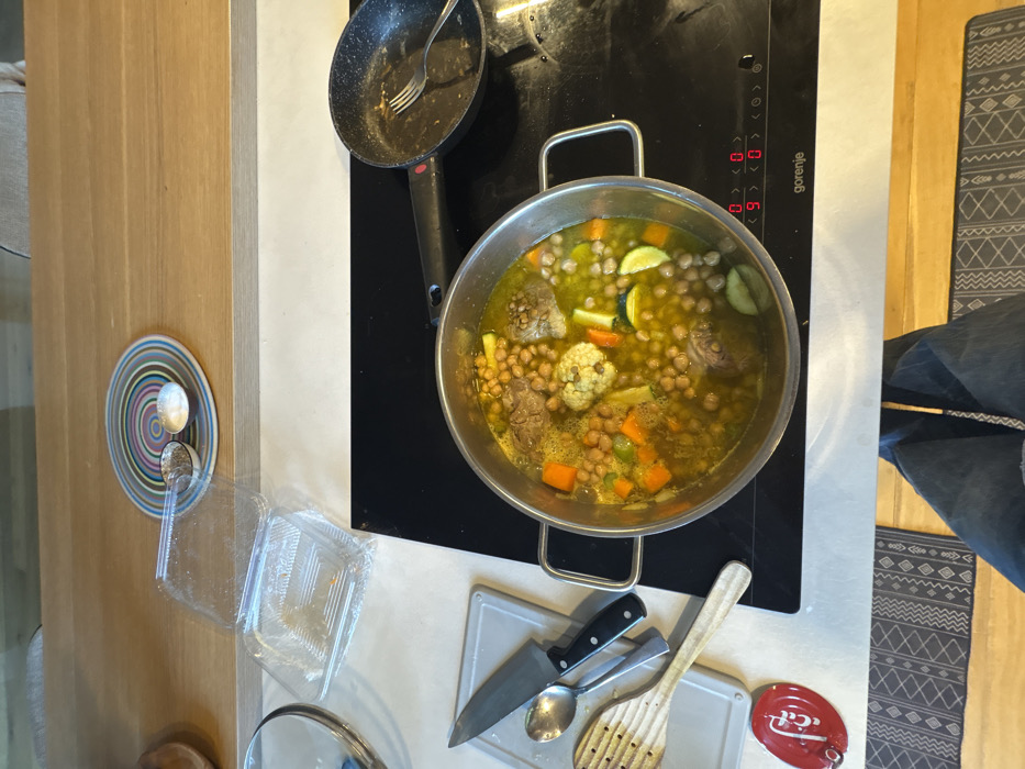 A top-down interior shot depicts a kitchen or dining area in Raanana, Israel. In the foreground, the lower legs of an individual wearing blue jeans are visible, positioned over a patterned grey rug on a light wooden floor. The midground features a white countertop and a black Gorenje induction cooktop. A large silver-colored pot containing a stew with chickpeas, meat, carrots, zucchini slices, and cauliflower florets in a yellowish broth is centrally positioned on the active cooktop, displaying a "9" and "0." To the right of the pot, an uncleaned black frying pan with food residue and a silver fork rests on the cooktop. To the left of the pot, on a light grey silicone mat, a chef's knife, a spoon, and a wooden slotted spoon are arranged. A partially visible red circular object, possibly a Coca-Cola branded item, is on the white counter below the utensils. An empty, clear plastic clamshell container is also on the white counter. In the background, a light wooden surface holds a round plate with concentric colored circles, upon which two small silver dishes contain dark and white granular substances, likely spices. A small portion of a person's light-colored attire is visible at the top edge of the frame, behind the wooden surface.