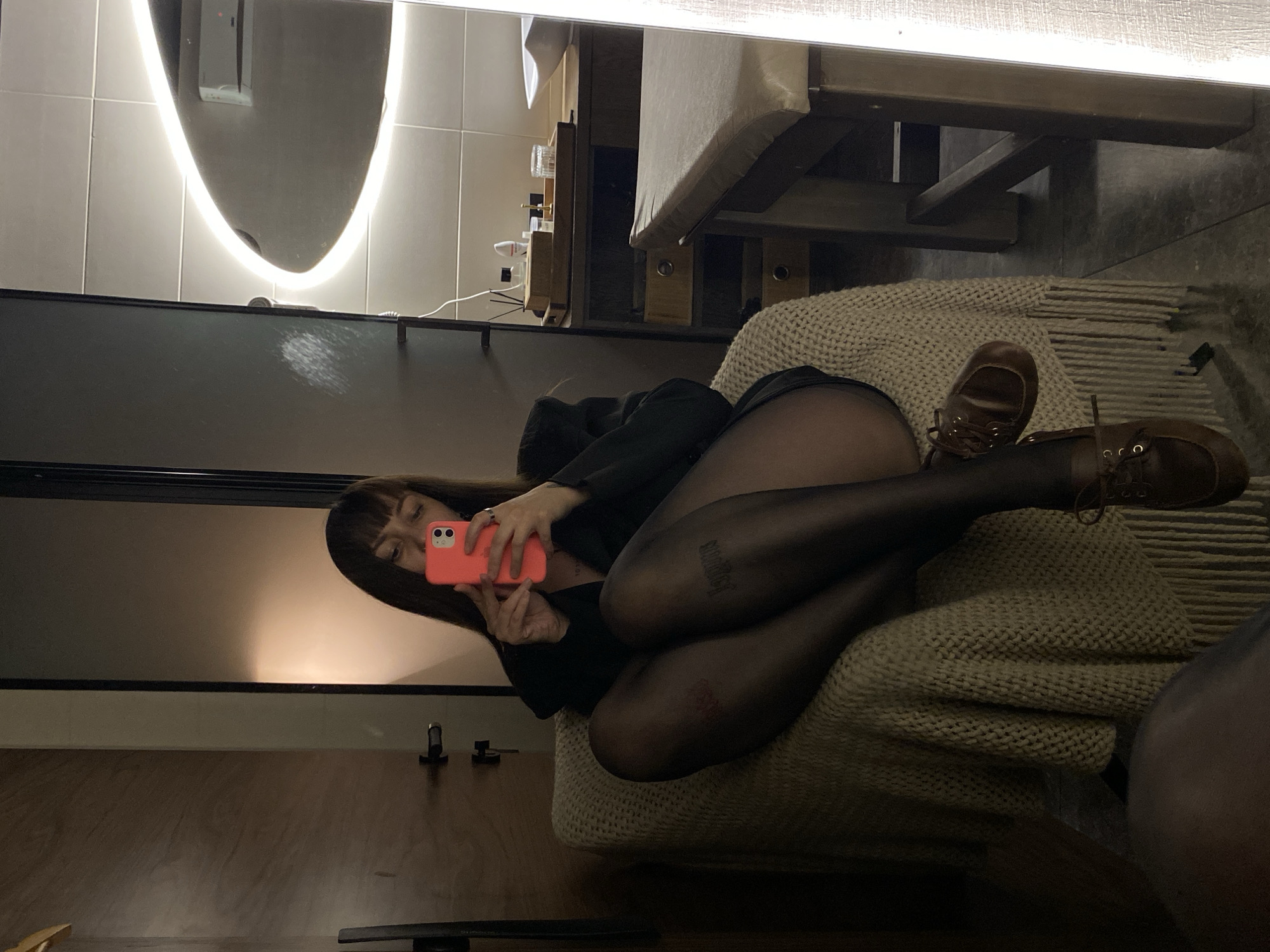 A woman is taking a selfie in front of a mirror. She is lying on a beige woven throw, wearing black tights, brown leather shoes, and a black top. The mirror reflects part of a room, including a curved lighted mirror and a wooden shelf with bottles. The location is likely indoors and cannot be confirmed as Votkinsk, Russia, based on the visual information provided.
