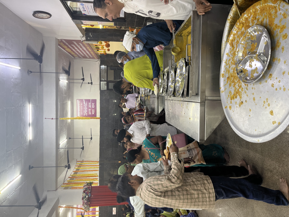 The image shows a line of people waiting to receive food at a langar, or free kitchen, in Panchkula, India. The coordinates provided are accurate and pinpoint the location to the Sector 26, Panchkula city. People are wearing a mix of modern and traditional clothing and are standing in line, some with plates in their hands. There are multiple serving dishes in the foreground, containing what appear to be rice, lentils, and vegetables. The image is taken from the perspective of someone standing to the left of the line, looking down the line toward the end of the langar. The overall atmosphere is peaceful and organized. The room is brightly lit, featuring multiple ceiling fans and several decorative garlands hanging from the ceiling. A sign on the wall reads "Aatma Dhan Maha Daan," which translates to "Gift of the Soul, Great Gift." It appears to be a place of worship or community service. The room is clean and well-maintained, and the people are moving in an orderly fashion. This suggests that the event is well organized and that the food is being distributed efficiently.