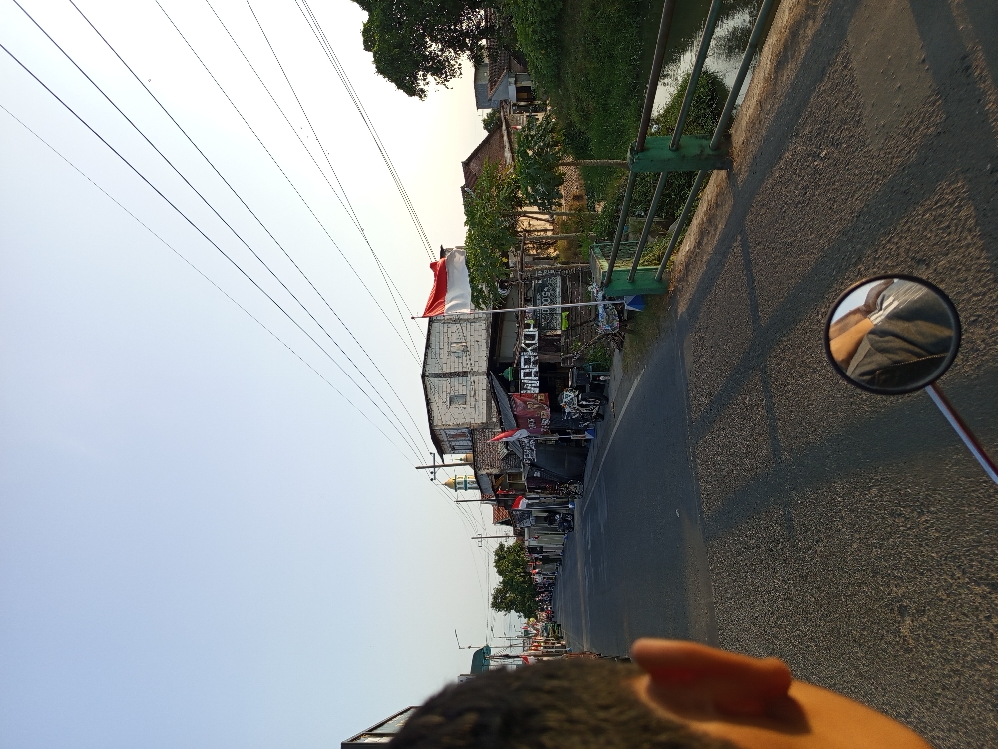 The image captures a street scene in Sumberejo, Indonesia, likely during the late afternoon or early evening, indicated by the warm, long shadows cast across the asphalt road. The perspective is from a moving vehicle, possibly a motorcycle, looking forward along the road. In the foreground, the back of a person's head and ear are visible on the left, and a motorcycle's rearview mirror is in the lower right, reflecting a person's shoulder and torso wearing a grey shirt.
The road itself is paved and appears to be a two-lane street with a slight incline. On the right side of the road, there is a green metal railing bordering a small canal or stream with lush green vegetation. Several trees are visible along the canal bank and further down the street.
The left side of the road features several buildings, some appearing to be small shops or warungs. Indonesian flags, red and white, are prominently displayed on poles outside some of these establishments, suggesting a national celebration or event. One building has a sign that appears to read "WARKO..." along with other text. A mosque with a green dome and minaret is visible in the distance. On the right side, further down the street, there are more residential-looking buildings with tile roofs.
The sky is clear and pale blue, transitioning to a lighter shade near the horizon, suggesting a sunset or sunrise. Power lines are strung across the sky, supported by poles along the street. Several motorcycles and a car are parked or moving along the road, indicating moderate activity. The overall impression is a typical Indonesian village or small town street setting.