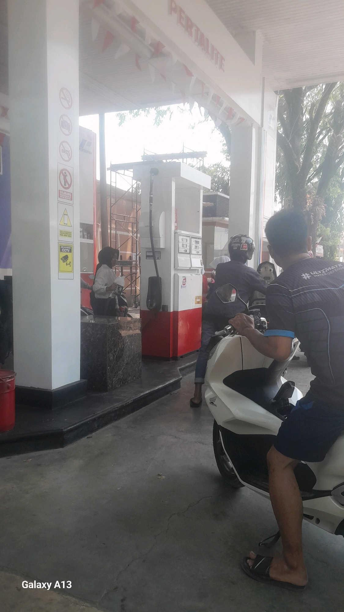 This is an outdoor, eye-level, medium shot photograph taken at a Pertamina gas station in Tanjunggemuk, Indonesia. In the foreground, a person on a white scooter is partially visible from the waist down, wearing dark blue shorts and black sandals. They are on the right side of the frame, facing left. To the left of the scooter, another person wearing a dark blue shirt with white lettering that appears to say "#ULTIMATE WATER DEFENSE" is seated on the scooter. The scooter's front is white.
In the mid-ground, behind the scooter, there is a red and white Pertamina gas pump. A person wearing a dark helmet and a dark blue t-shirt is seated on a scooter facing the gas pump. They are holding a white container, possibly a gas canister. To the left of the gas pump, a woman is seated on a scooter, wearing a light-colored shirt and dark pants. She is holding a white container with a red label.
The background shows construction scaffolding and a building. The area is paved with concrete. Overhead, the words "PERTALITE" are visible on a white structure, along with red and white pennant flags. The lighting suggests it is daytime, with some dappled sunlight. The lower left corner of the image contains the text "Galaxy A13".