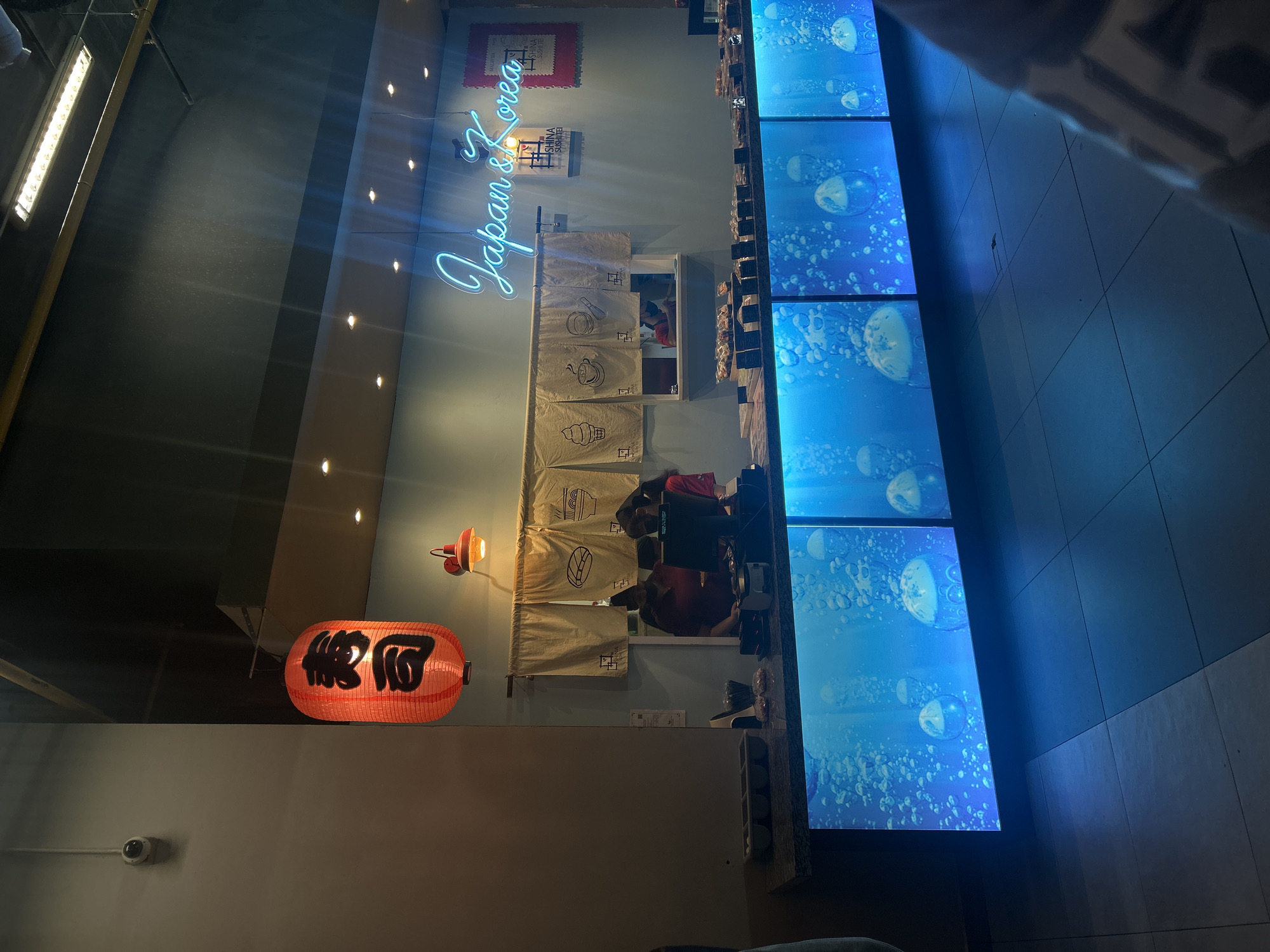 A vertically-oriented, slightly rotated image captures the interior of a restaurant or eatery. The establishment's name, "Japan & Korea," is displayed in blue neon lettering near the upper portion of the frame. Below the sign, there are small decorative items on the wall. A red Japanese paper lantern with black Kanji characters is suspended to the left. The interior features people seated at tables, suggesting diners or staff. A large, multi-panel display to the right shows an animated image of bubbles. The restaurant appears to be in Kuala Lumpur, Malaysia based on the restaurant name and architectural style.