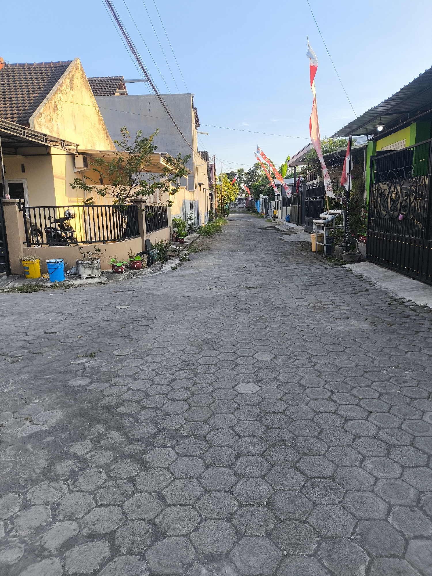 This is a street-level view of a residential alley in Lumajang, Indonesia, likely during daytime, with the sun casting warm light on the left side of the scene. The alley is paved with hexagonal concrete blocks. On the left, there are houses with tiled roofs and light-colored walls. One house has a black metal fence in front of it, with a motorcycle parked behind it. Several colorful buckets, including yellow and blue, are placed near the fence. A small tree grows next to the house. Further down the alley, on both sides, are more houses and gates. Red and white Indonesian flags are hung along the street, suggesting a national celebration or event. The sky is clear and blue.