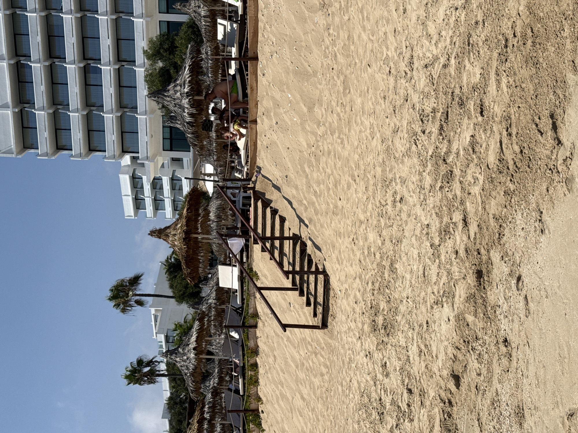 A sunny day at Nissi Beach in Ayia Napa, Cyprus, is depicted. The foreground is dominated by a vast expanse of golden sand, textured with footprints and small pebbles. A set of wooden stairs with metal railings ascends from the sand, leading to a raised beach area. This area is furnished with numerous straw umbrellas providing shade for white sun loungers. In the background, a modern white hotel building with multiple balconies stands prominently against a clear blue sky. Several palm trees are scattered across the scene, adding to the tropical ambiance. A group of people are visible relaxing on sun loungers under one of the umbrellas.