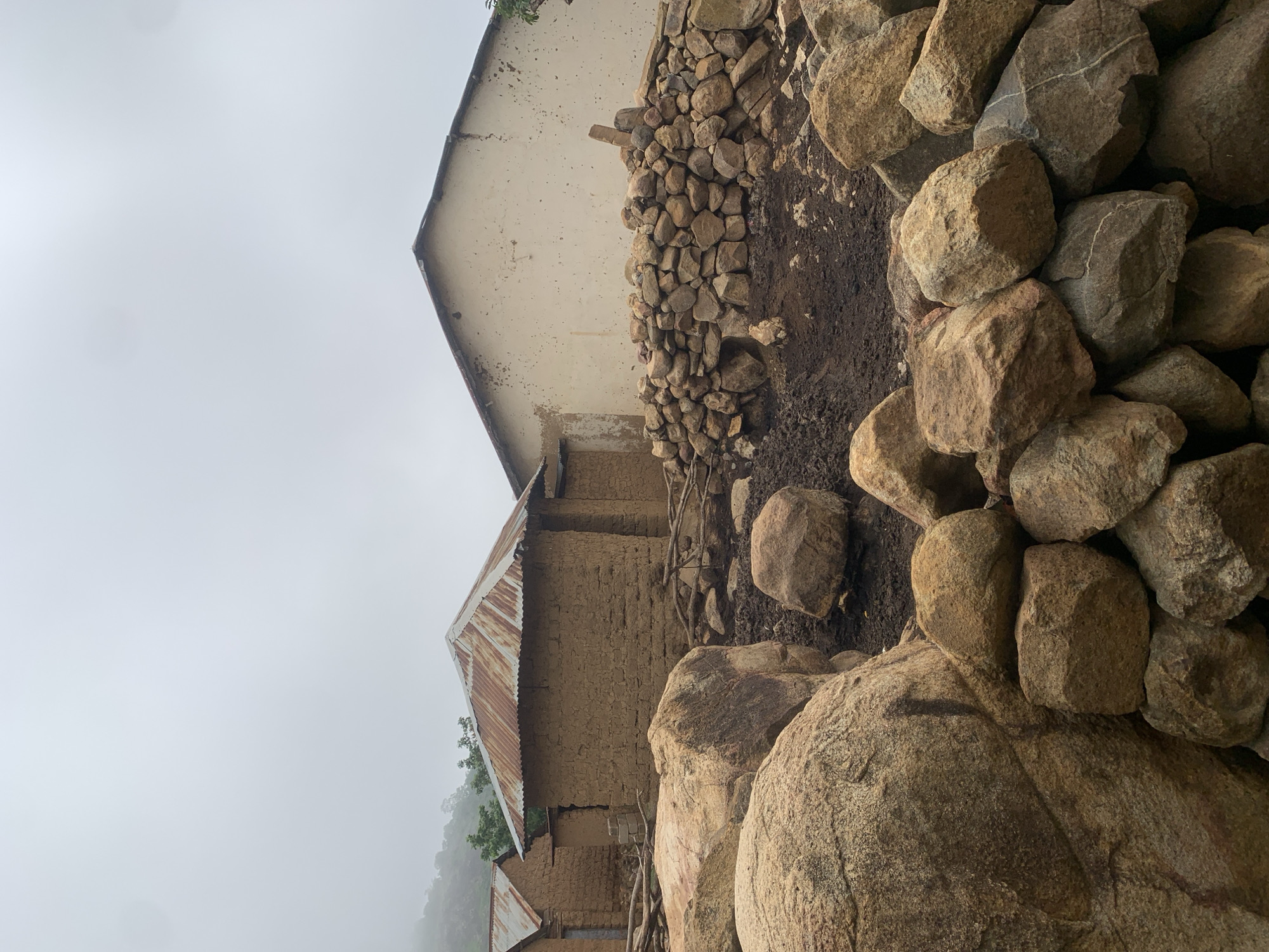 The image shows a collection of traditional mud brick houses with corrugated metal roofs in Zhwarki Mile 10, Nigeria. The houses appear to be old and weathered, with some showing signs of disrepair. In the foreground, a pile of large rocks and a wall constructed from smaller stones are visible. The environment is overcast, with a hazy sky and hints of greenery on a distant hillside. The scene conveys a sense of rural simplicity and perhaps hardship. There are no people visible in the image.