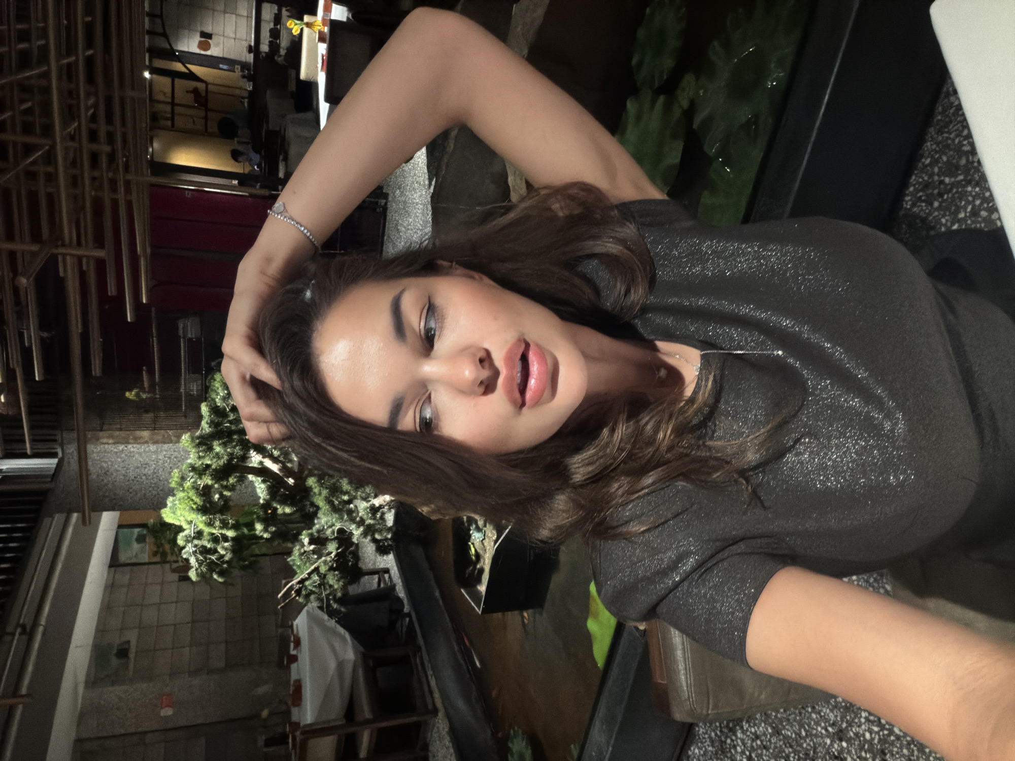 A woman is posing for a selfie indoors in Dongcheng District, China. She wears a sparkling black top and a bracelet on her left wrist. Her dark brown hair is wavy. She holds her left hand on her head. In the background, there is a decorative green plant and what appears to be outdoor restaurant seating. The location is a dark interior of a restaurant, likely at night.