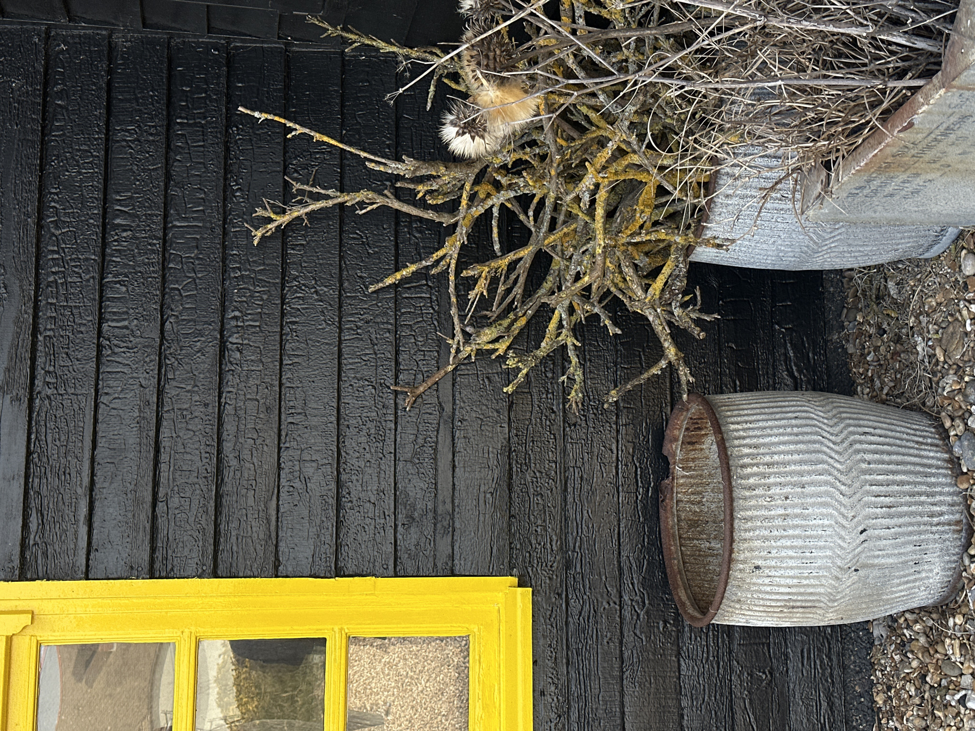 The image depicts a close-up view of a building's exterior. The wall is clad in charred black timber planks. A yellow window frame is visible in the lower left corner, featuring what looks like glass panes with obscured views inside. To the right of the wall, two old metal tubs rest, one partially filled with dry twigs and dried seedheads. The tubs are corrugated. The ground around the tubs appears to be covered with small pebbles. The location is inferred to be in the Folkestone and Hythe District, United Kingdom based on the metadata.
