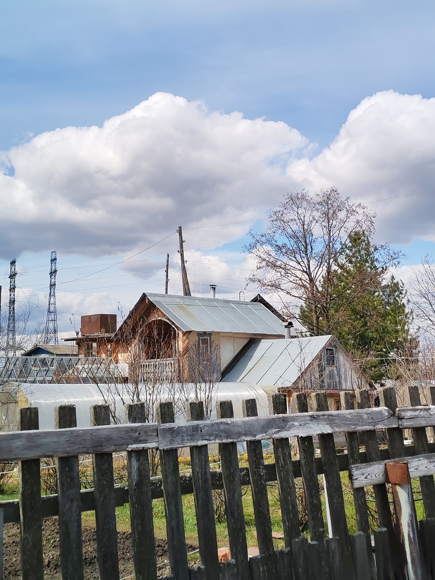 The image captures a rural or semi-rural landscape, likely in the Chaikovsky region of Russia, on a bright, partly cloudy day. In the immediate foreground, a weathered, rustic wooden picket fence stretches across the lower half of the frame, its vertical slats and horizontal rails showing signs of age. Behind the fence, patches of green grass and bare earth are visible.
The middle ground features several buildings and natural elements. On the left, two long, low, translucent structures, likely greenhouses, are present. Further back, multiple wooden houses with contrasting silver metal roofs stand out. One prominent house boasts an ornate wooden facade with a covered porch and detailed carvings. Another smaller structure with a similar roof is partially visible to its right. Scattered throughout the scene are trees, including a tall, bare deciduous tree suggesting early spring or late autumn, and several darker evergreen trees.
In the background, particularly on the left, tall metal lattice towers, indicative of high-voltage power lines, rise prominently against the sky. Wooden utility poles and a network of overhead wires also traverse the scene.
The sky is a vivid blue, adorned with large, fluffy white cumulus clouds, suggesting fair weather. The scene depicts a quiet, static environment with no visible human activity or text.