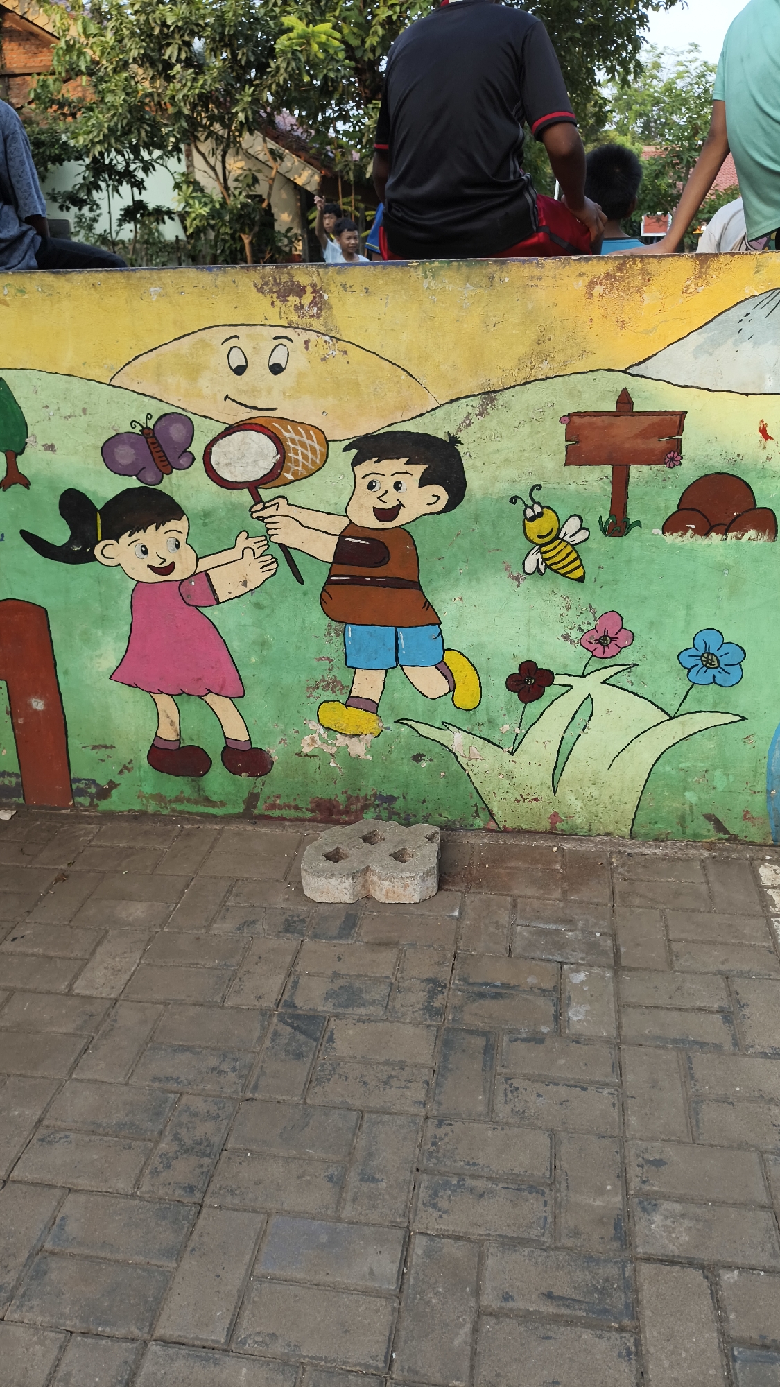 The image shows a mural painted on a wall, depicting two children playing with a butterfly net. A boy in a brown shirt and blue shorts holds the net, while a girl in a pink dress reaches out towards it. A butterfly is shown near the girl. In the background, there is a sun, a bee, flowers, a signpost, and two mounds that appear to be rocks or dirt. The mural is painted on a weathered wall with some peeling paint. The ground in front of the wall is paved with interlocking bricks. In the upper portion of the image, out-of-focus people are visible sitting and standing on top of the wall, with trees and buildings in the background. The scene is likely located in a public outdoor area in South Jakarta, Indonesia, given the visual cues of the environment.