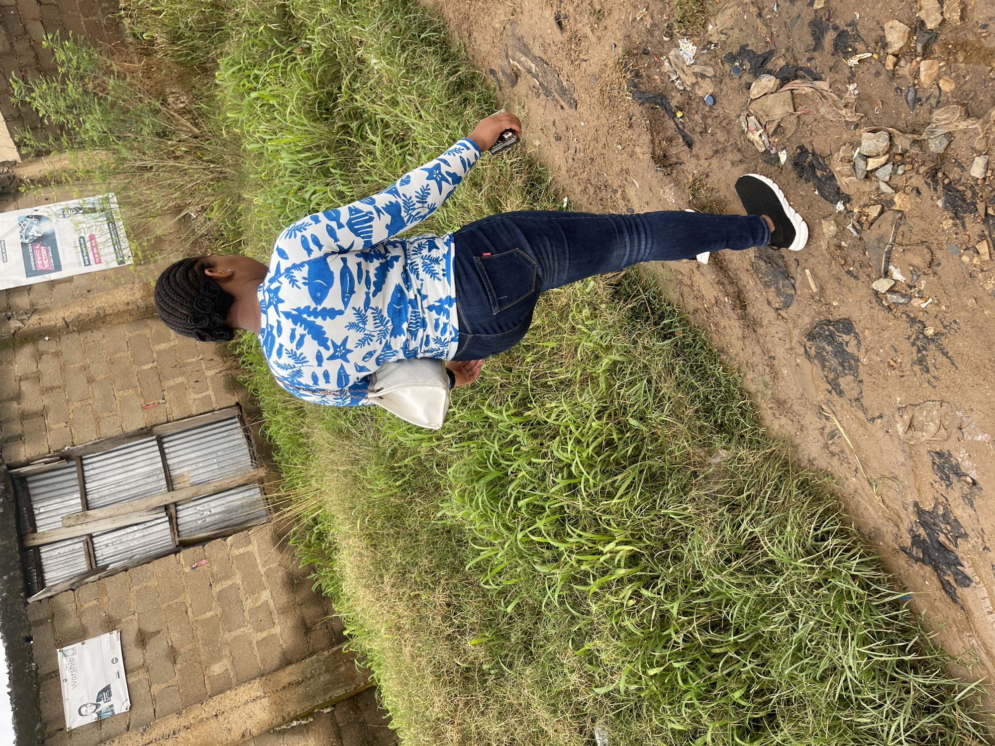 This full-body shot captures a Black woman from behind, standing in an outdoor setting that appears to be Rafin Par, Nigeria. She is wearing a long-sleeved blue and white fish-patterned shirt, dark blue jeans, and black and white slip-on sneakers. A white purse is slung over her left shoulder. Her hair is braided and styled into a bun at the back of her head. She is holding a small object, possibly a phone or camera, in her right hand.
The woman is standing on a path that transitions from green grass to a dirt and gravel area strewn with debris, including plastic bags and rocks. To her left, a low-rise building constructed of concrete blocks is visible, with corrugated metal covering openings that resemble windows. A poster is affixed to the wall of the building. The scene suggests an everyday moment, possibly a walk or pause during an outdoor excursion. The lighting and greenery indicate it is daytime, with no clear indicators of specific weather conditions. The overall impression is one of a casual, unposed moment in a semi-urban or rural environment.
Visible text on a poster on the building reads "Worship" in large letters. Other text is present but not clearly legible at this resolution.