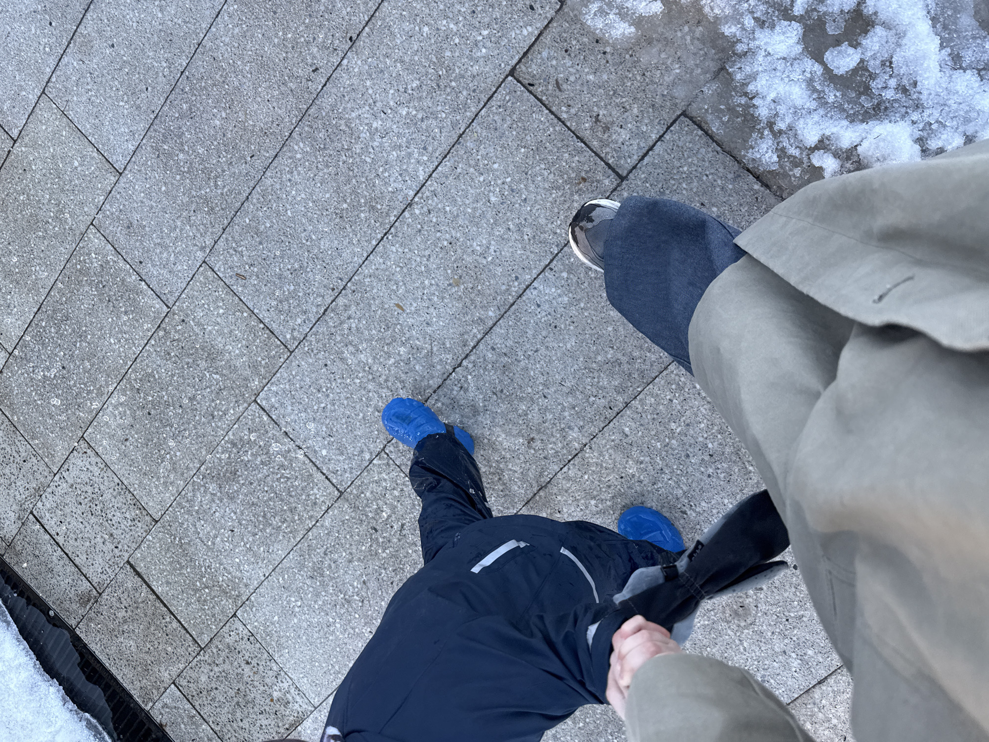 The image shows a first-person perspective of two people standing on a paved surface, likely a sidewalk or pedestrian area. The person taking the picture is wearing a beige trench coat and blue jeans with sneakers. Next to them is a child wearing a dark blue snowsuit and bright blue overshoes. The child is holding a dark grey umbrella and holding hands with the adult. Some snow and ice are visible near the edge of the pavement. The location is described as Chaikovsky, Russia.