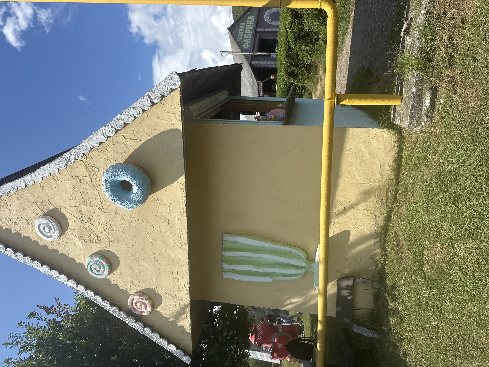 The image depicts a whimsical, gingerbread-house-themed structure with a yellow facade, adorned with decorative candy-like motifs and a striped design. The structure appears to be a small stand or shop, with an open window suggesting it serves customers. In the background, a building with Cyrillic lettering that reads "ЧАЙНАЯ ФАБРИКА" (Tea Factory) is visible, indicating a commercial establishment. Lush green foliage and a clear blue sky with scattered clouds frame the scene. A yellow pipe runs across the lower portion of the image, adding a utilitarian element to the otherwise fantastical setting. The overall environment suggests an outdoor recreational or tourist area.