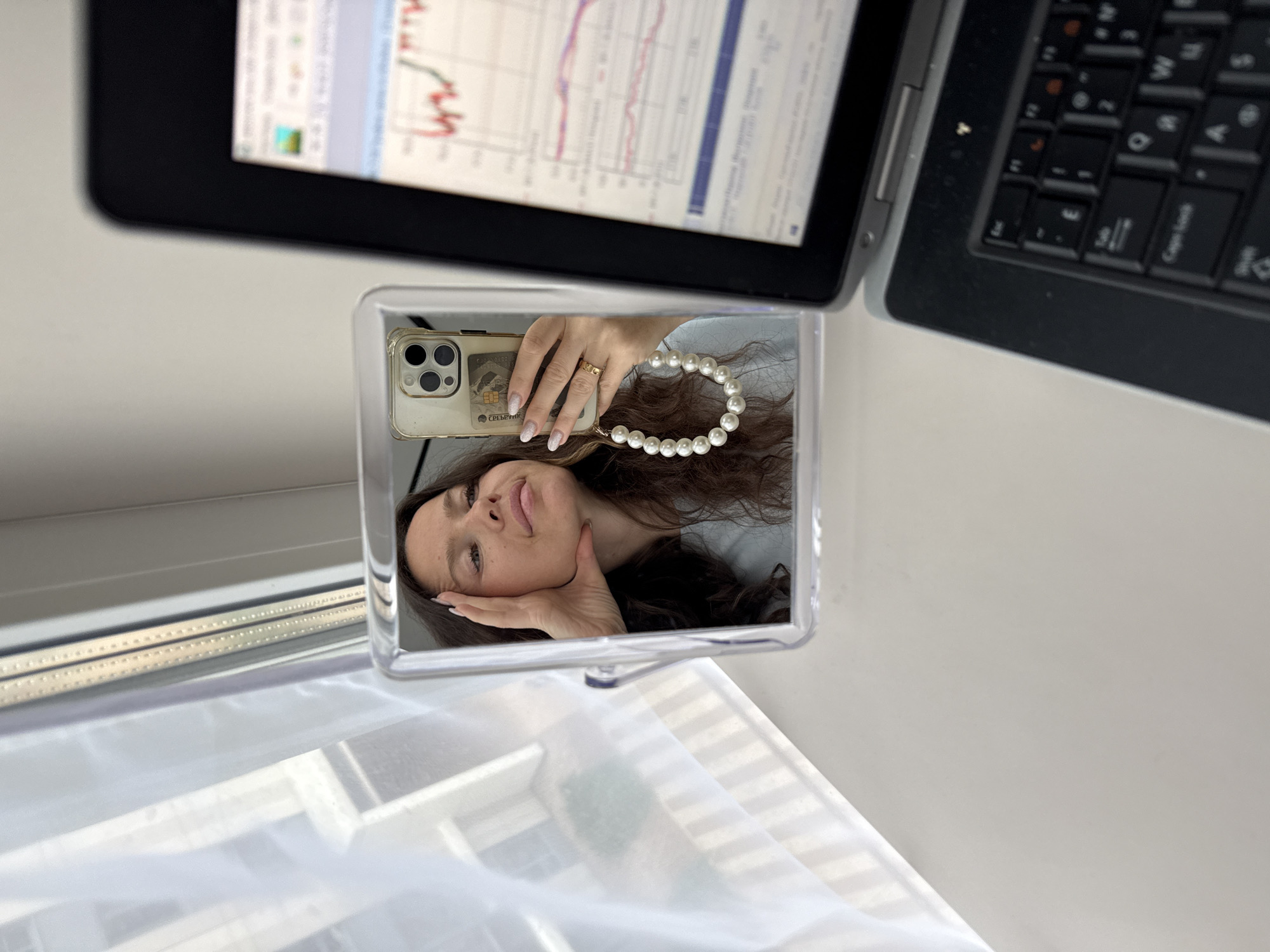 The image shows a close-up of a woman taking a selfie in a mirror. She has long, dark, wavy hair and is wearing a light blue shirt. Her left hand is resting on her cheek, and she is wearing a ring on her right ring finger. Her phone, which is in a clear case with a pearl strap and a card inserted, is visible in the mirror. A laptop with a financial chart displayed on the screen is in the foreground, to the right of the mirror. The scene appears to be indoors, near a window with sheer white curtains. The overall impression is that of a workspace or study area.