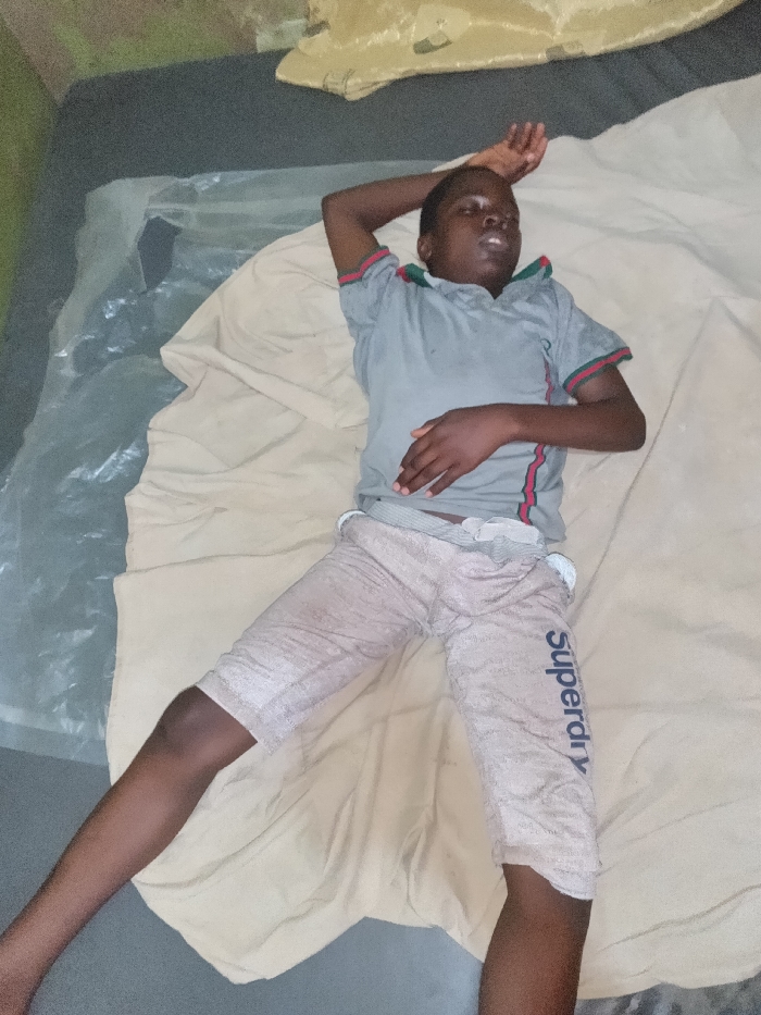 The scene depicts a single male juvenile or young adult lying supine on a sleeping surface within an indoor environment in Onitsha, Nigeria. The individual, with dark skin and short dark hair, is positioned centrally in the frame. His eyes are partially open, and his mouth is slightly ajar. He wears a grey short-sleeved polo shirt with red and green striped accents on the collar and sleeve cuffs. Light-colored, knee-length shorts, marked with "Superdry" printed vertically on the outside of the right thigh, are worn, with a light-colored undergarment or waistband visible above them.
The individual's right arm is bent, with the hand resting on his forehead. His left arm is also bent, with the hand resting on his lower abdomen. He lies on a light-colored fabric sheet, which partially covers a transparent plastic sheet. Both layers rest on a dark, flat surface, likely a mattress or floor. The immediate background includes a section of a plain greenish wall in the upper left and a crumpled, light-colored fabric or object above the individual's head. The lighting is ambient, without a discernible direct source, suggesting an interior room. No other individuals or active interactions are present.