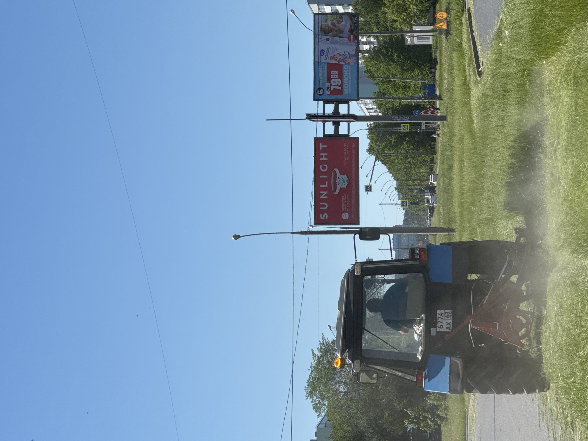 The image shows a tractor mowing a grassy area beside a road in an urban environment. The tractor is black and blue with the license plate "6774 AX 63". A person is visible inside the tractor's cabin. The mown grass is being ejected from the side of the tractor.
In the background, there are billboards advertising "SUNLIGHT" and "МОЛОКО". There are trees lining the street and buildings in the distance. There are street lights, power lines, and road signs visible.