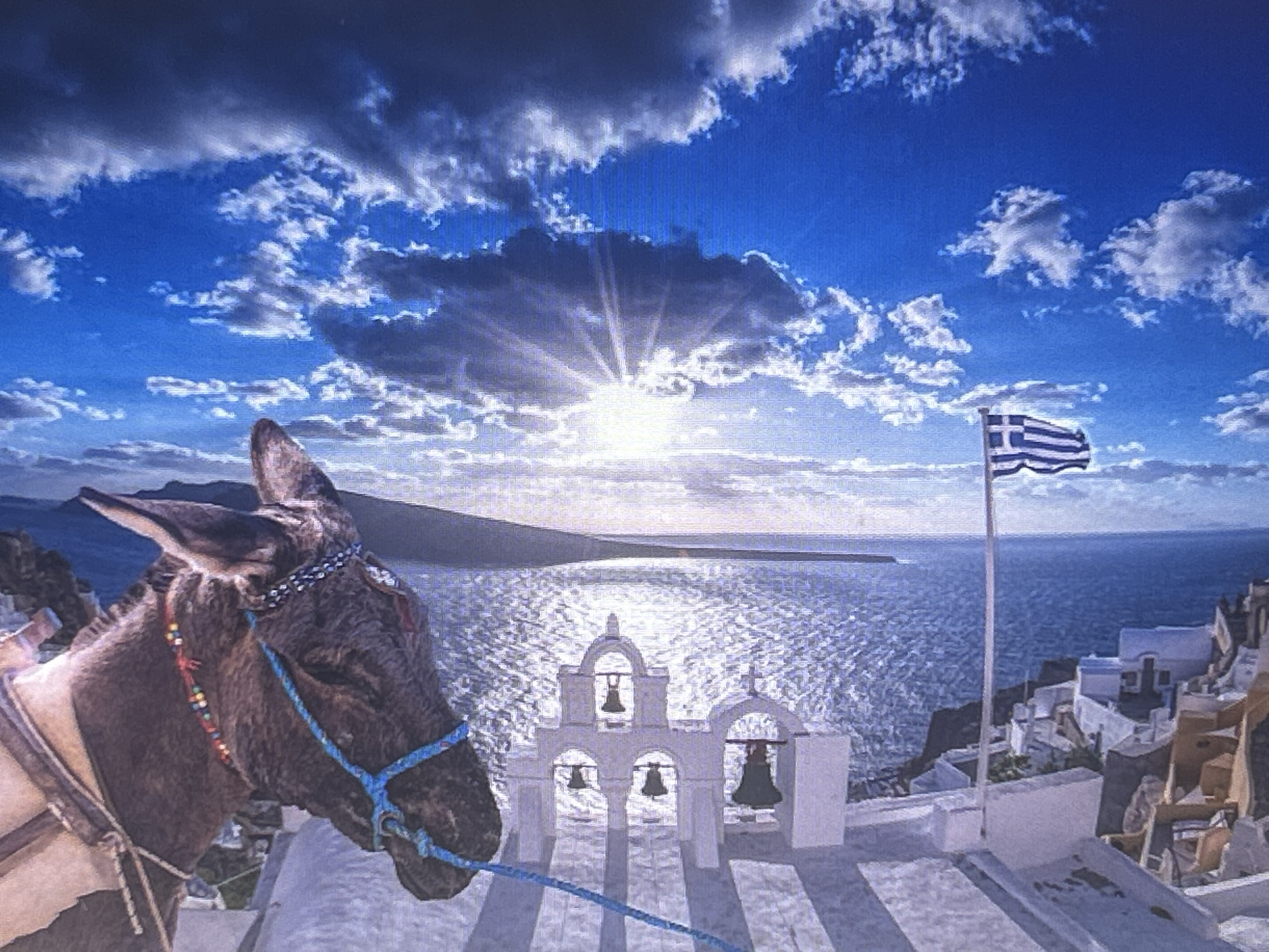 A donkey stands in the foreground of a picturesque scene, its head and left side filling the left portion of the frame. It wears a decorative bridle with beads and a blue rope. In the background, a white church with multiple bells and arched openings stands on a white surface. Beyond the church, the sparkling blue sea stretches to the horizon. A Greek flag waves on a flagpole to the right of the church. The sun is setting or rising, casting brilliant rays through scattered clouds, creating a dramatic effect in the blue sky. The white buildings of a village are visible on the cliffside to the right. The overall impression is one of a serene and beautiful location. The image appears to be a photograph with an artistic filter applied, giving it a painterly quality. There is no visible text. The location context provided is Sofia, Bulgaria, which is incongruous with the visual elements of the image, such as the Greek flag and the architecture strongly suggesting a Greek island like Santorini.
