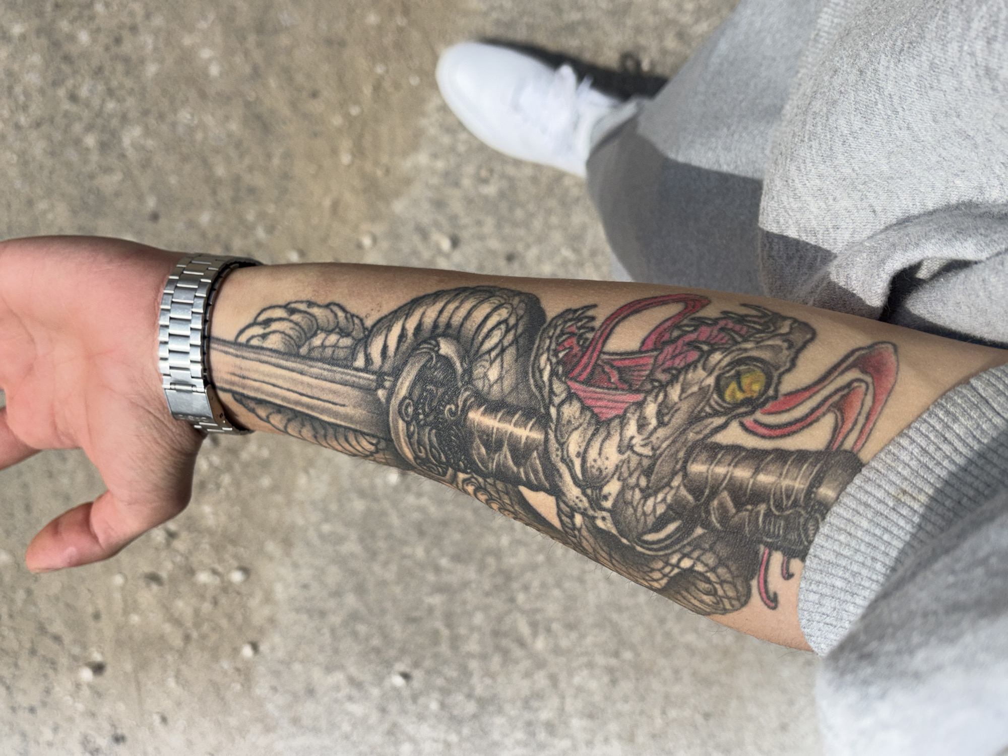A close-up, eye-level shot features an arm with a prominent tattoo of a snake coiled around a sword. The snake has a visible eye and red accents. The arm is adorned with a silver-colored wristwatch. The person is wearing a grey sweatshirt or hoodie. In the background, a glimpse of a white sneaker suggests the person is standing on a textured, grey surface that appears to be concrete or asphalt. The context does not include information regarding location or Bulgaria.