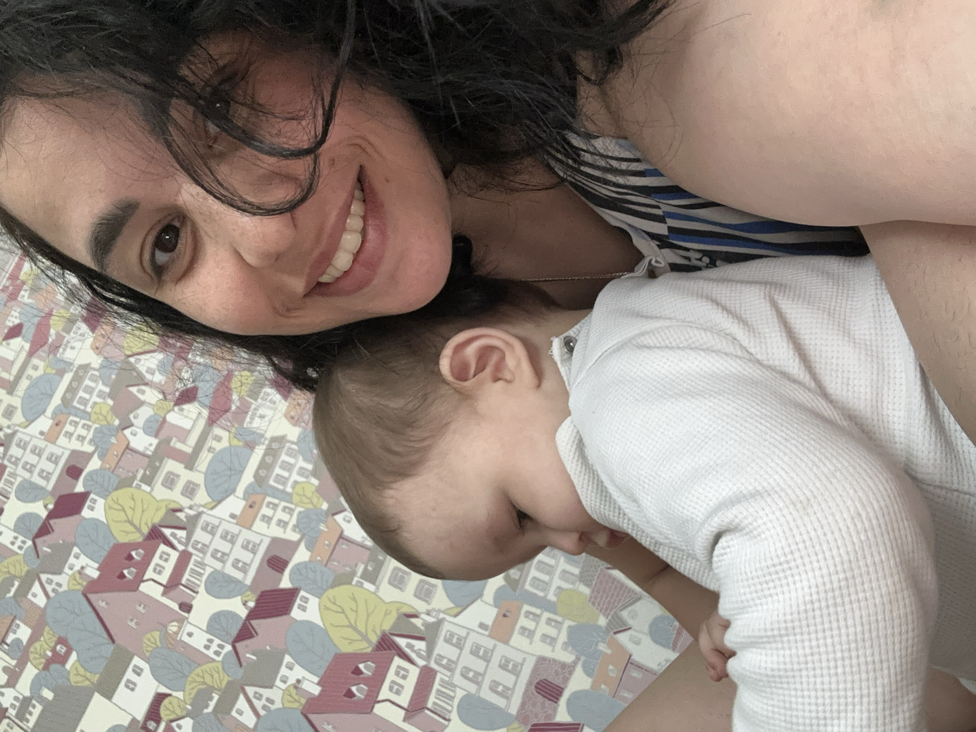 An overhead selfie captures a woman and a baby in an intimate, indoor setting. The image is tilted, with the woman positioned in the upper left, smiling broadly directly at the camera. She has dark, curly hair, brown eyes, and is wearing a blue and white striped top. Her expression is warm and happy.
Beside and slightly below her, a baby is visible, looking down and to the right, away from the camera. Only the baby's head, ear, and part of the upper body are clearly seen. The baby has light brown hair and is dressed in a white, waffle-knit long-sleeved outfit.
The background features a whimsical pattern of cartoon-like houses, trees, and shrubs in shades of pink, white, gray, and yellow on a light background. This pattern covers the surface beneath them, possibly a bedspread, rug, or wallpaper. The overall mood is tender and joyful, highlighting a close moment between parent and child. No text is visible in the image.