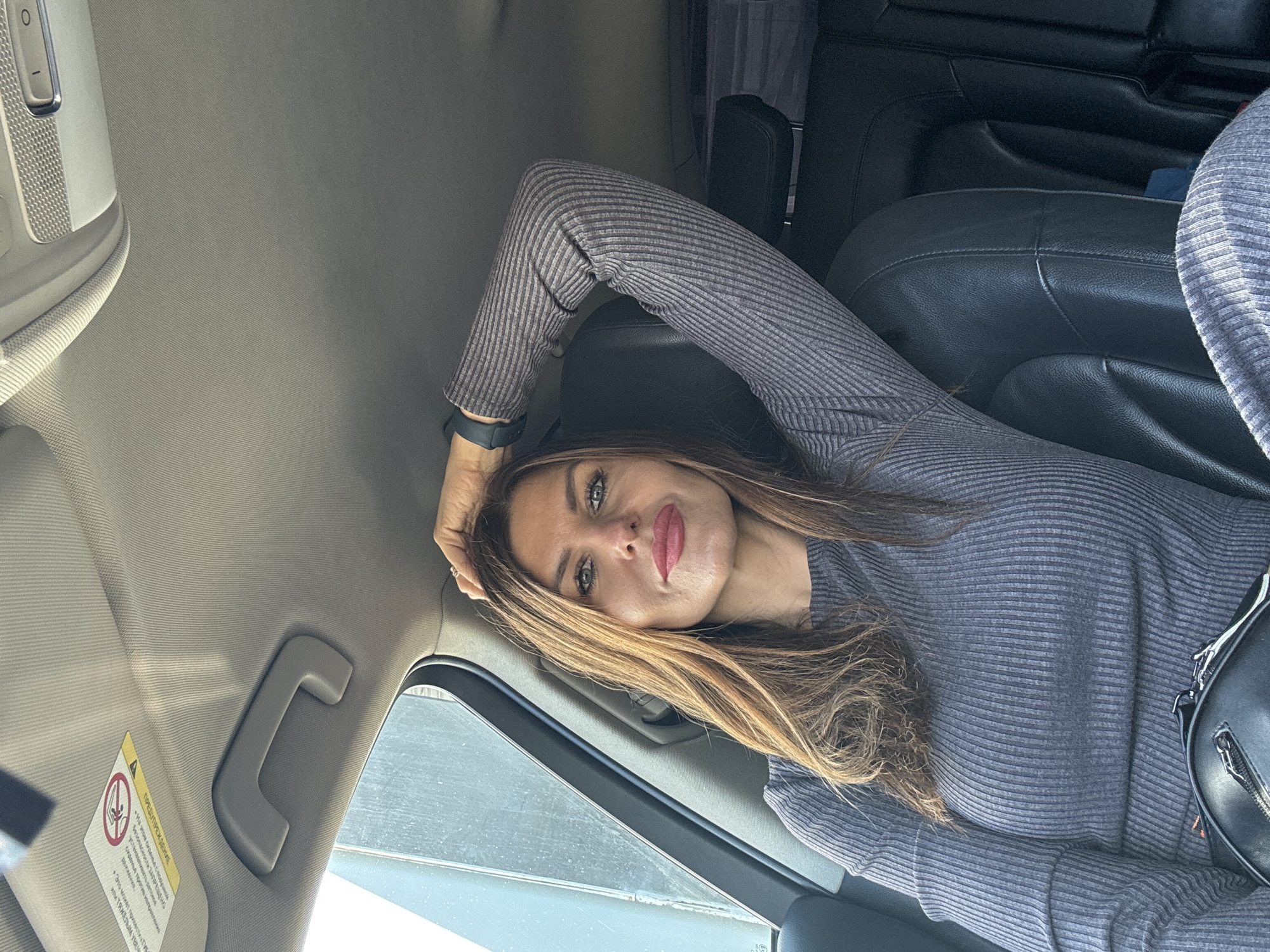 A woman is lying inside a car. She has long, light brown hair and is wearing a gray, long-sleeved shirt. Her head is near the car window, and her arm is bent, with her hand resting near the car's interior roof. Black leather seats are visible. The location is Chaikovsky, Russia, based on the text on the warning sticker in the car.