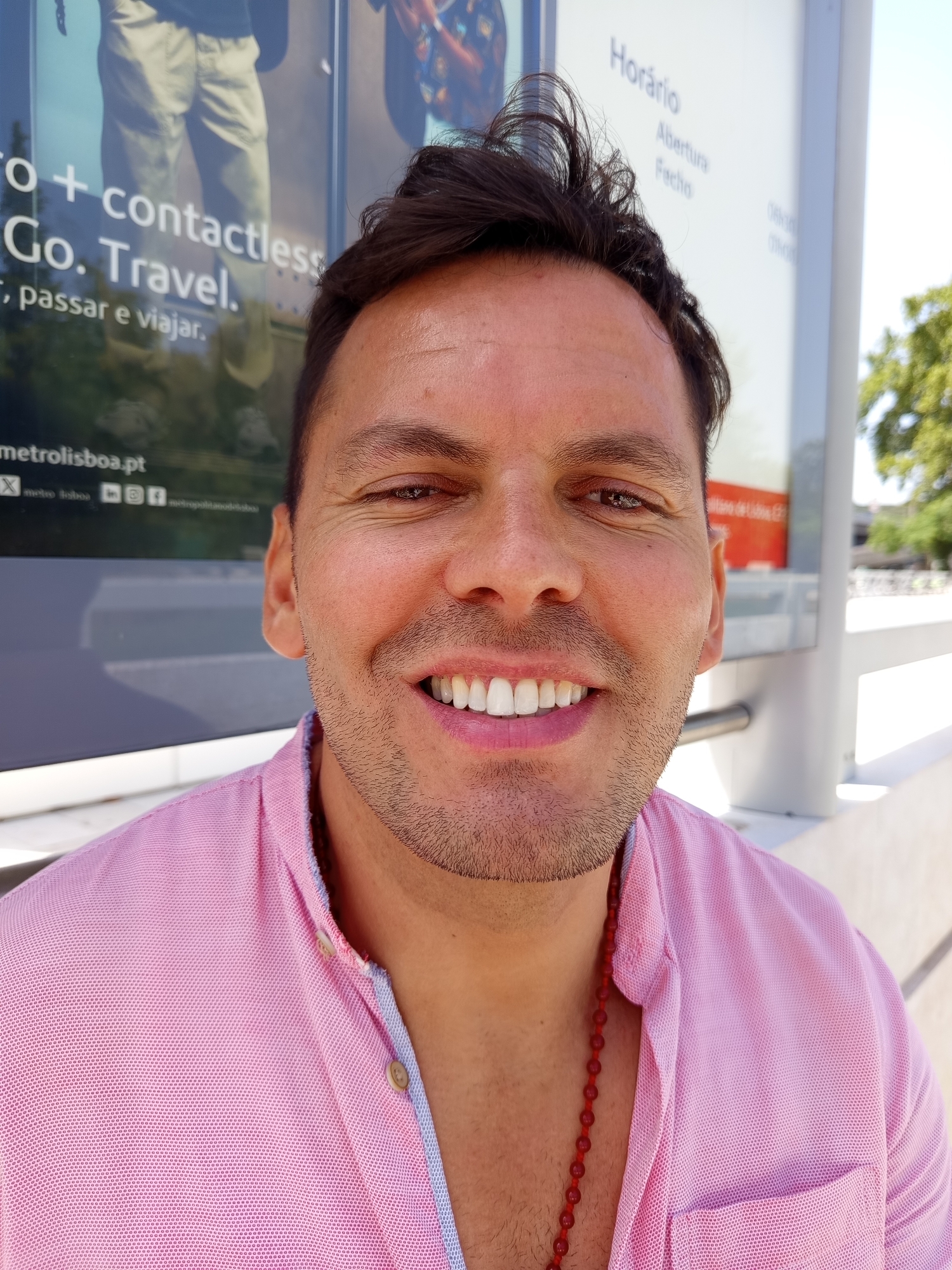 The image is a close-up portrait of a man with medium-toned skin, short dark hair, and a light stubble beard. He is wearing a pink collared shirt with light blue trim and a red beaded necklace. He is smiling, showing white teeth, and looking towards the camera. In the background, there are posters with text, including the word "Horário," which is Portuguese for "schedule" or "hours." The posters also display information about "metro lisboa." The presence of "metro lisboa" indicates that the man is likely at a metro station in Lisbon, Portugal. The background also shows a bright sky and some trees.