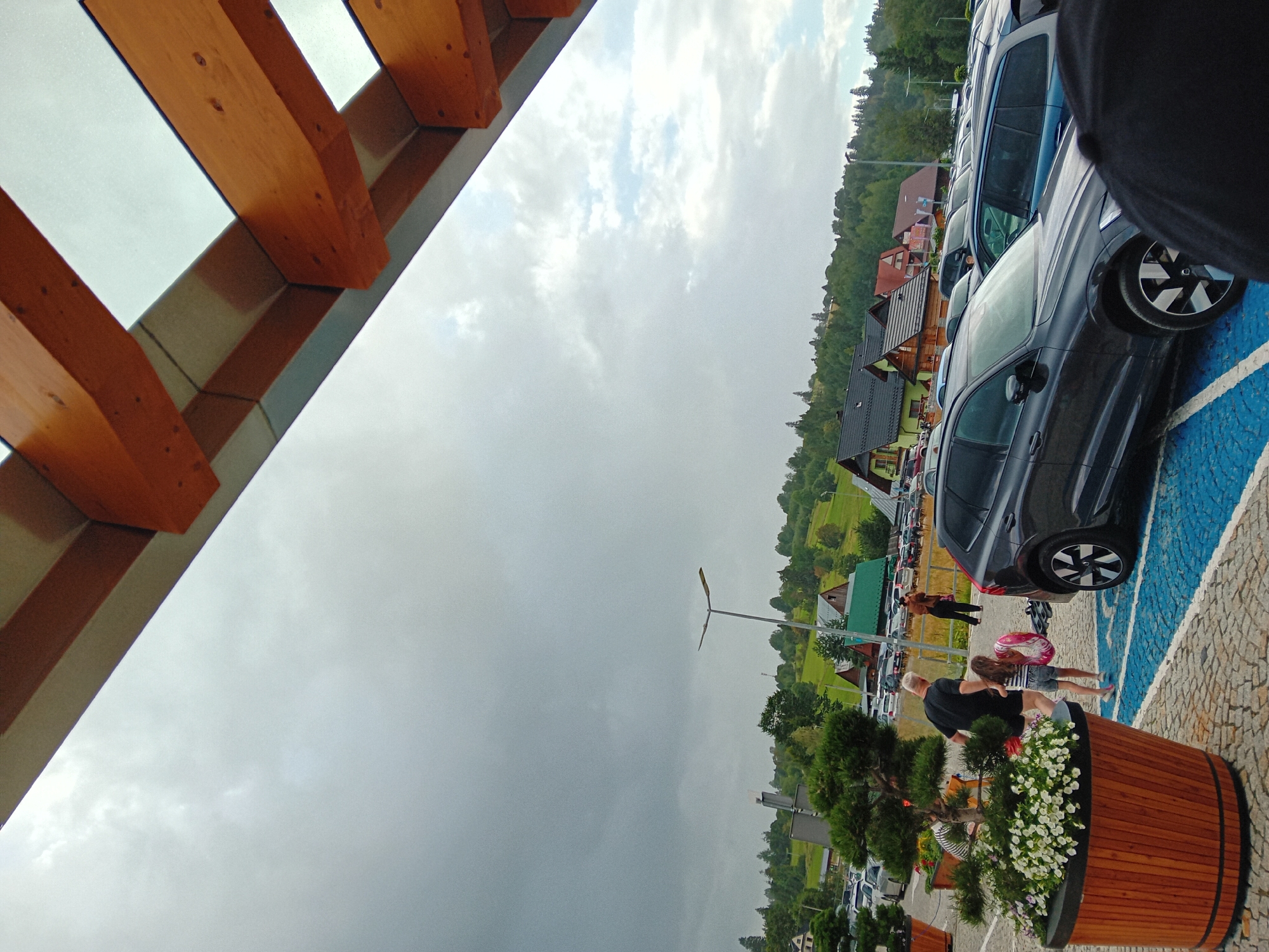 The image captures a cloudy day in Chochołów, Poland, with a view of a parking lot and a mountainous landscape. In the foreground, a large wooden planter filled with white flowers is visible. To the right of the planter, a dark grey SUV is parked. A man and a young girl are walking in the parking lot, with the girl holding a pink inflatable ring. In the background, numerous cars are parked, and traditional wooden houses with dark roofs are nestled on a green, tree-covered hillside. A streetlamp stands tall in the middle ground. The sky is overcast, with a mix of dark and lighter clouds. The architecture of the houses suggests a rural or semi-rural setting within the Chochołów region, known for its preserved folk architecture.