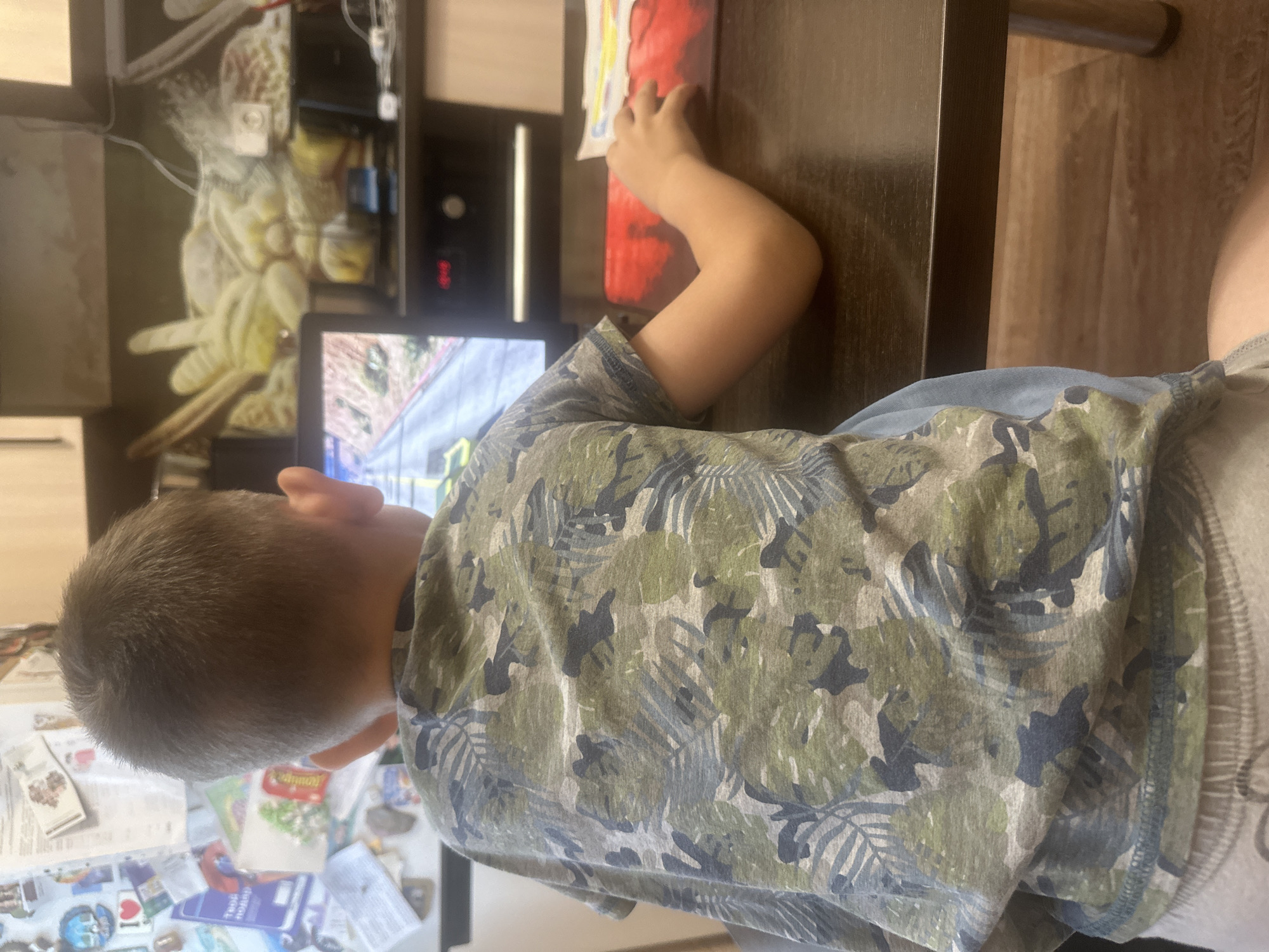 A young child, viewed from behind, sits at a dark wooden table playing a video game on a tablet. The child wears a camouflage t-shirt and light-colored shorts. The tablet screen displays a racing game. To the right of the tablet, a hand is visible resting on a red mousepad. In the background, a kitchen area is partially visible, with shelves containing various items, including what appears to be a mural of a tropical fruit. A refrigerator, covered in magnets and papers, is to the left of the frame. The floor is made of light-colored laminate. The scene is indoors, likely in a home kitchen in Novoaltaysk, Russia.
