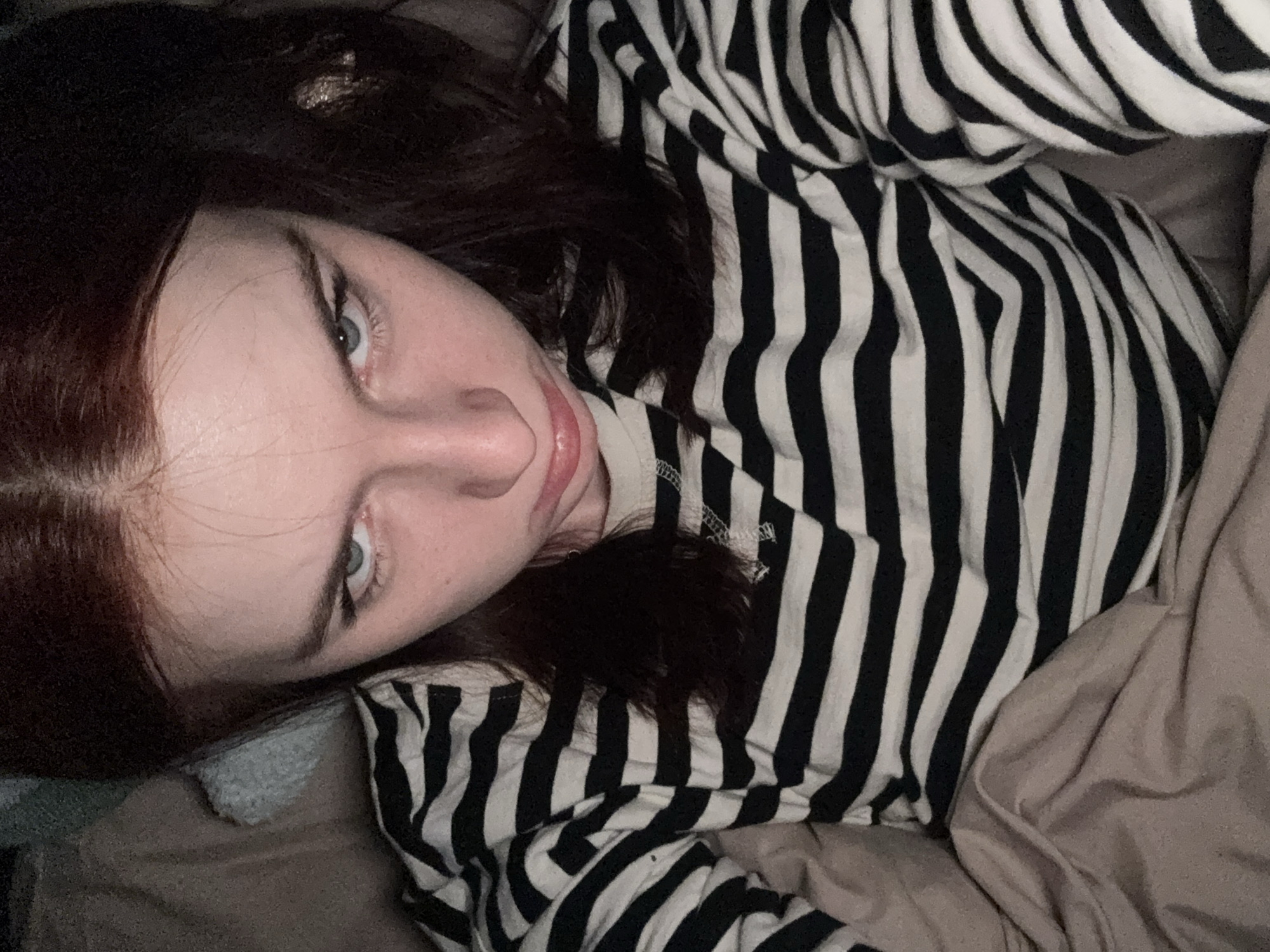 A person lies on a tan bed with a grey pillow, wearing a black and white striped shirt. The person has dark hair with some auburn highlights and light-colored eyes, and is looking at the camera. The location is unspecified, and there's no indication of Saint Petersburg, Russia.