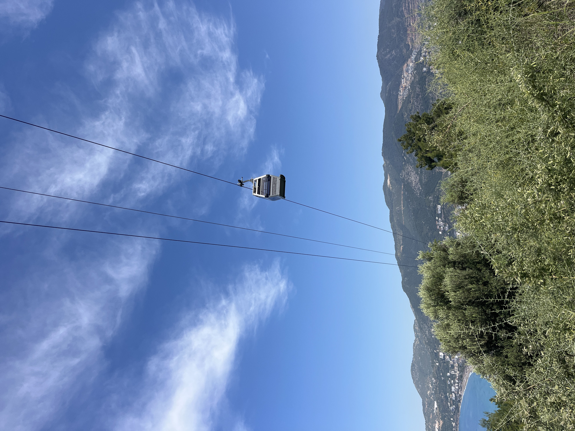 The image features a cable car suspended in mid-air, against a backdrop of a partially cloudy, blue sky on the left and mountains with greenery and buildings on the right. The cable car is light-colored, and its carriage shape is visible. The vegetation is dense and a coastline with a beach and a small town can be glimpsed in the lower right corner. The location is likely in Alanya, Turkey, based on the landscape and architectural context. The picture shows the cable car going up a mountainside near the beach of a coastal city.