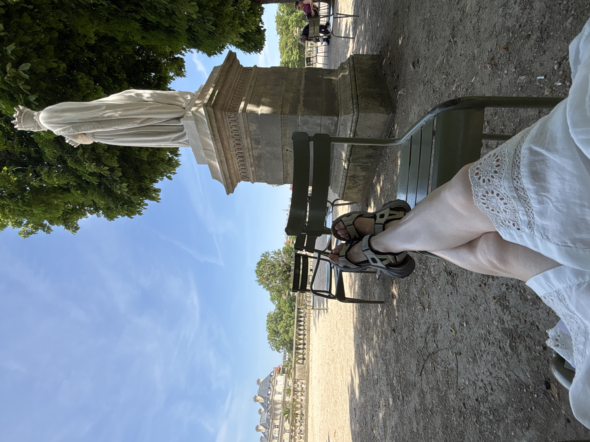 The image shows a close-up of a person's legs, wearing a white dress and sandals, as they sit on a green metal park bench. The person's legs are crossed, and the white dress has a lace trim along the hem. In the background, a stone statue on a pedestal is visible, surrounded by lush green trees. The ground is covered in gravel, with shadows cast by the trees, suggesting it is daytime. Further in the background, there are more park benches and people walking, and a building with classical architecture, consistent with the appearance of a Parisian park or garden. The sky is a clear blue with a few wispy clouds. The overall scene suggests a relaxed and pleasant atmosphere in an outdoor setting, likely during the day. There is no visible text in the image.