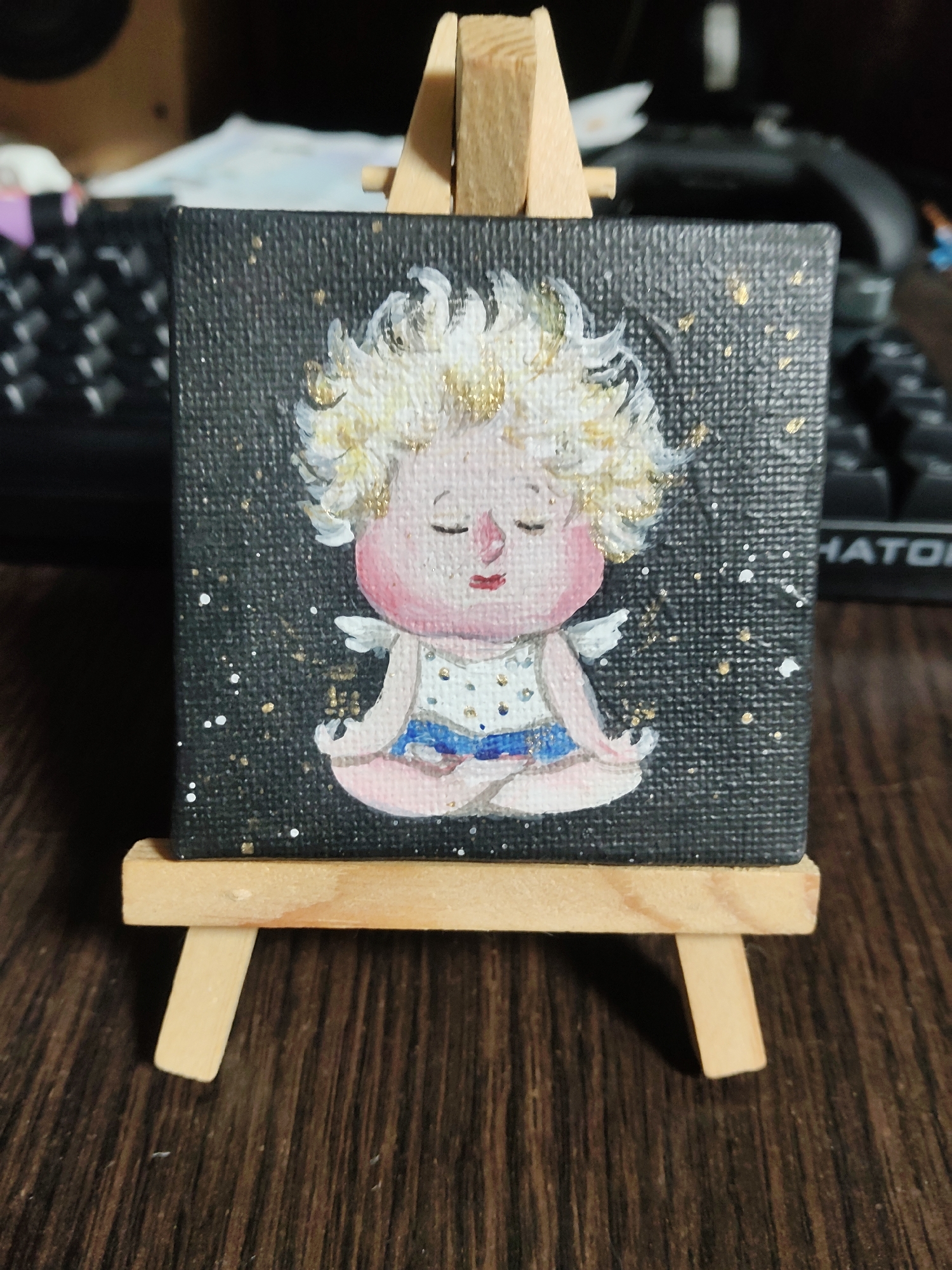 A miniature painting of a cherubic figure meditating is displayed on a small wooden easel. The figure has fair skin, closed eyes, and a halo of golden, curly hair. It is adorned with a white top speckled with gold and a blue sash. Tiny white wings are visible on its back. The background of the painting is black and speckled with white and gold dots, resembling a starry night sky. The easel is made of light-colored wood and rests on a dark, wooden surface, possibly a desk. In the blurred background, a black keyboard and a wooden speaker are partially visible. The lighting is soft, highlighting the details of the painting and its display.