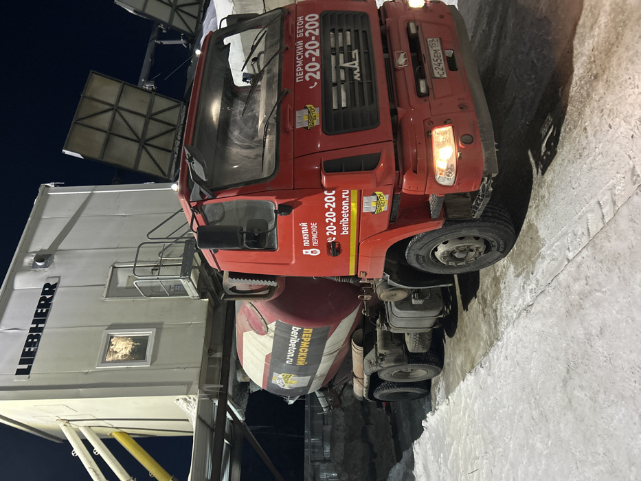 An illuminated industrial scene in Залесная, Russia, captured at night, features a red MAZ concrete mixer truck parked on a snow and ice-covered surface. The truck's cabin and mixing drum are emblazoned with "ПЕРМСКИЙ БЕТОН," "20-20-200," and "beribeton.ru." Its headlights and right-side turn signal are active, and the Russian license plate reads C245EM 159. Positioned above the truck is an elevated white industrial module, likely a concrete batching plant, bearing the "LIEBHERR" brand name. This structure includes a visible illuminated window, a door with a small access platform, and associated industrial piping, including a prominent yellow chute to the left. No individuals are present, and the scene is lit by artificial sources against a dark night sky.