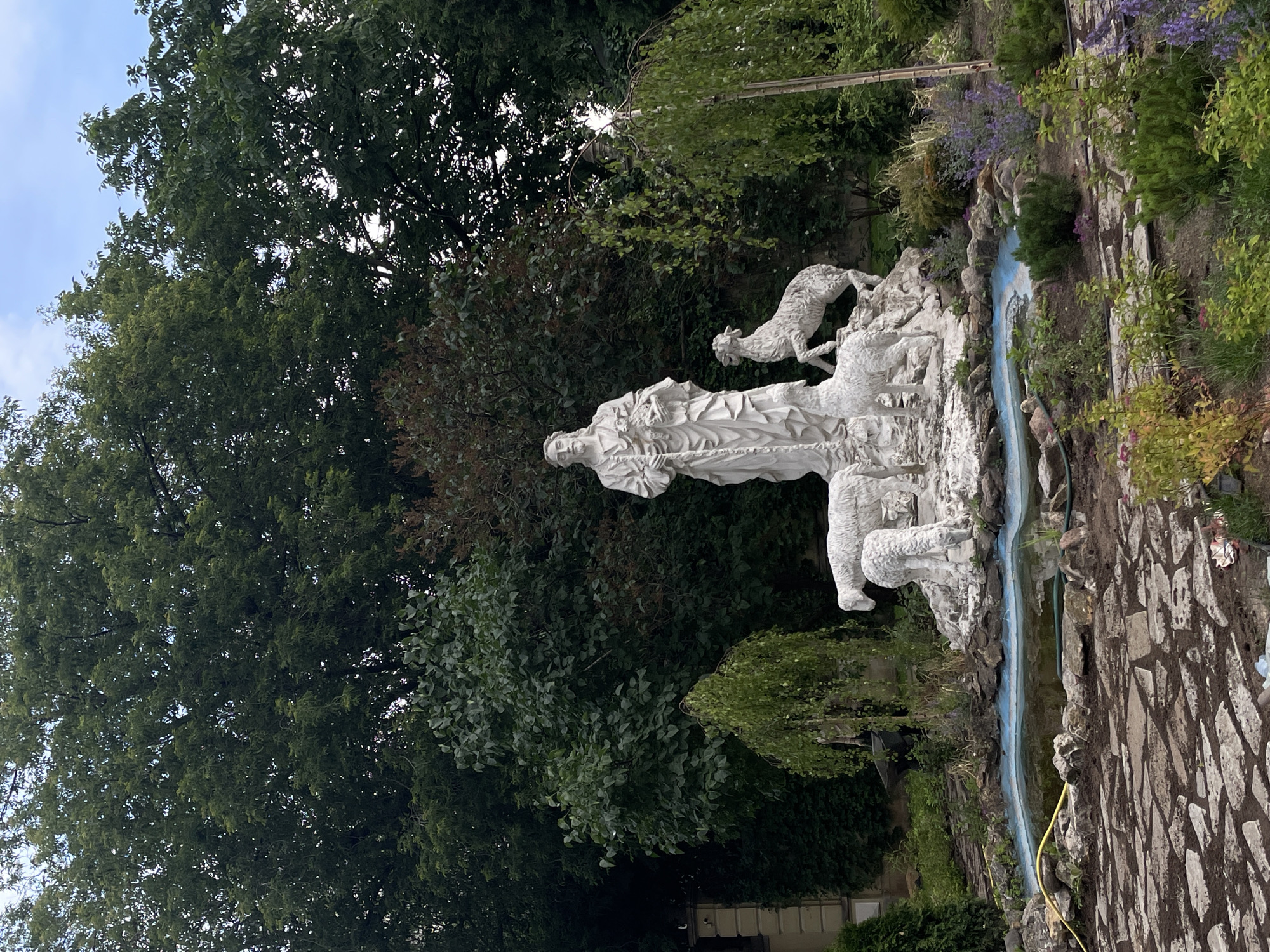 The image shows a white stone sculpture of Jesus Christ as the Good Shepherd, holding a lamb and surrounded by three other sheep. The sculpture is set in a garden with a small pond in the foreground and lush green trees in the background. The area around the pond is paved with irregular flagstones, and there are various plants and shrubs. The sculpture appears to be an outdoor installation, possibly in a park or a religious institution. The specific location in Moscow, Russia is not identifiable from the image. The scene is static, with no observable activity or interaction between subjects.