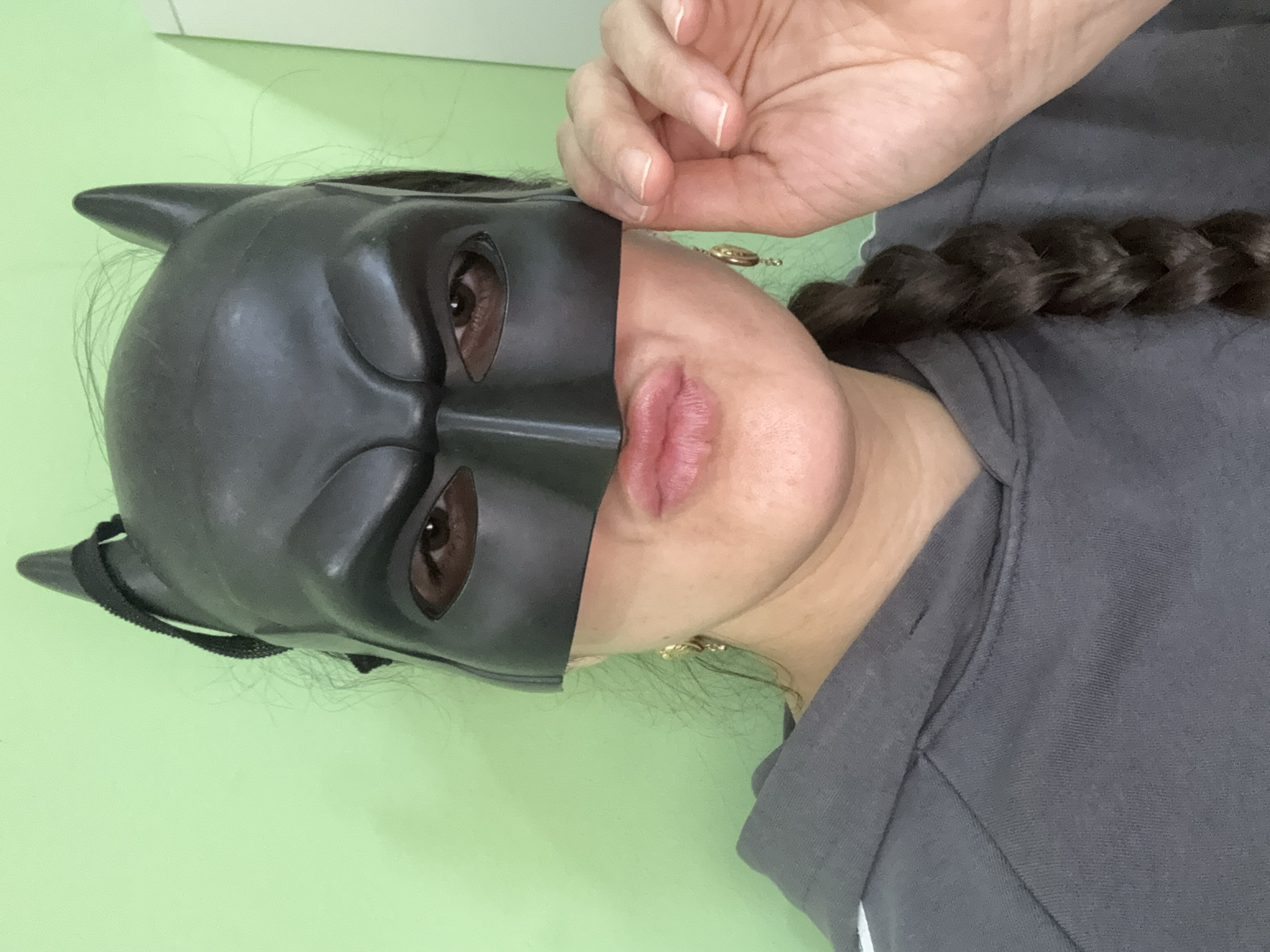 A close-up, head-and-shoulders portrait depicts a person, likely a woman, indoors in Tobolsk, Russia. The subject's left half of the face is obscured by a black, rigid Batman mask with pointed ears, through which the left eye is visible. The right half of the face is uncovered, showcasing the nose, slightly parted pink lips, and cheek. The visible eye has a neutral expression.
Dark brown braided hair rests on the subject's right shoulder and chest, with small strands escaping from under the mask. A hand with manicured, natural nails is positioned in the upper right quadrant of the image, appearing to hold or adjust the mask near the subject's temple. The person is wearing a gray hooded sweatshirt, visible on the right shoulder and chest, which features a subtle white stripe detail. A small, golden, circular earring or charm is visible near the right earlobe. The background is a plain, light green wall, indicating an uncluttered indoor setting. The image captures a playful or casual moment with the costume accessory. No text is visible in the image.