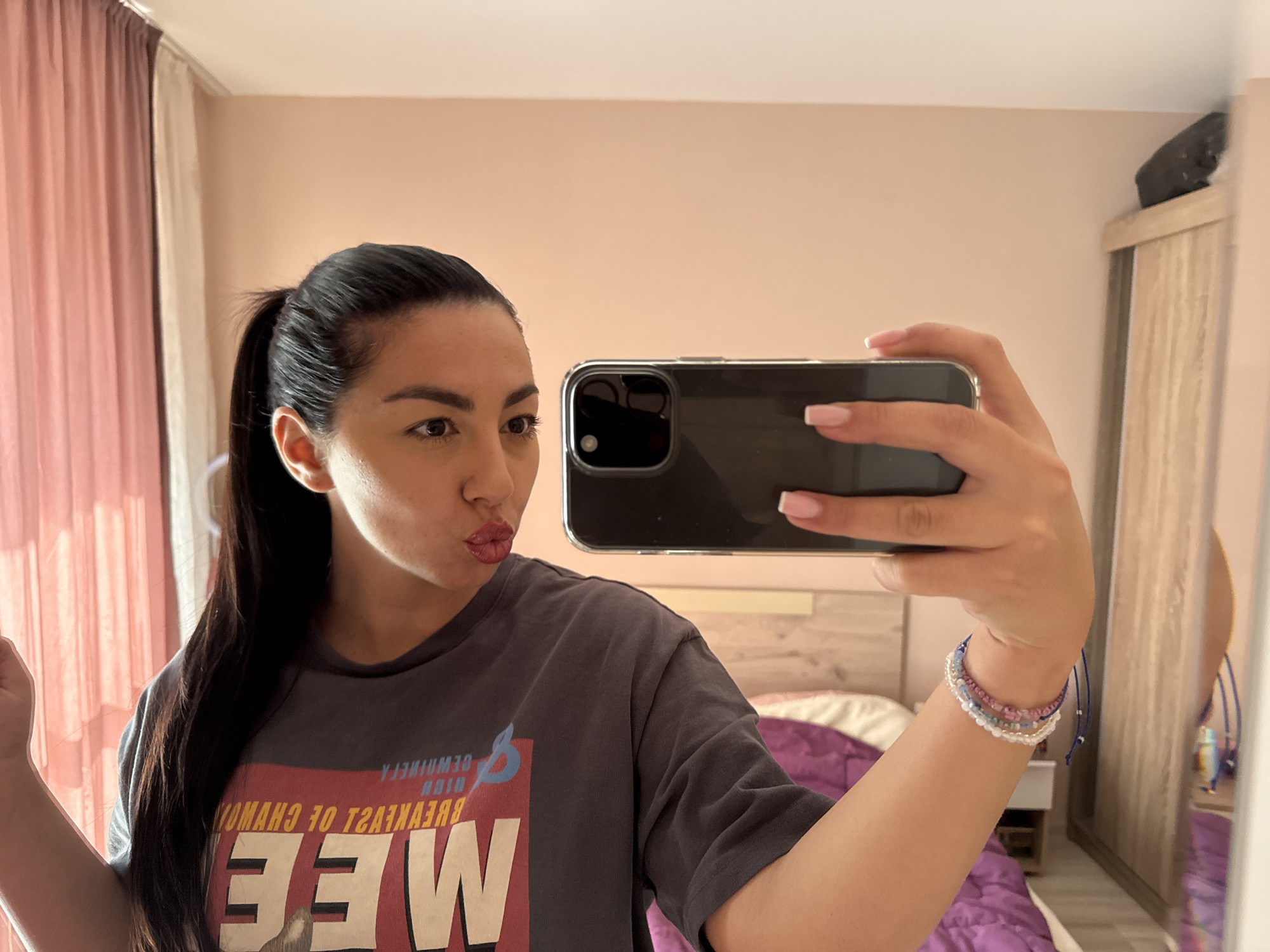 A woman is taking a selfie in what appears to be a bedroom. She has dark hair pulled back in a ponytail, and is wearing a gray t-shirt with a graphic design on the front. She is making a kissing face. She is holding a smartphone up with her right hand, which has a bracelet on the wrist. The room has pale pink walls, a light-colored wooden bed with a purple blanket, curtains, and a wardrobe in the background. It is impossible to verify the location as Pernik, Bulgaria, without additional information.