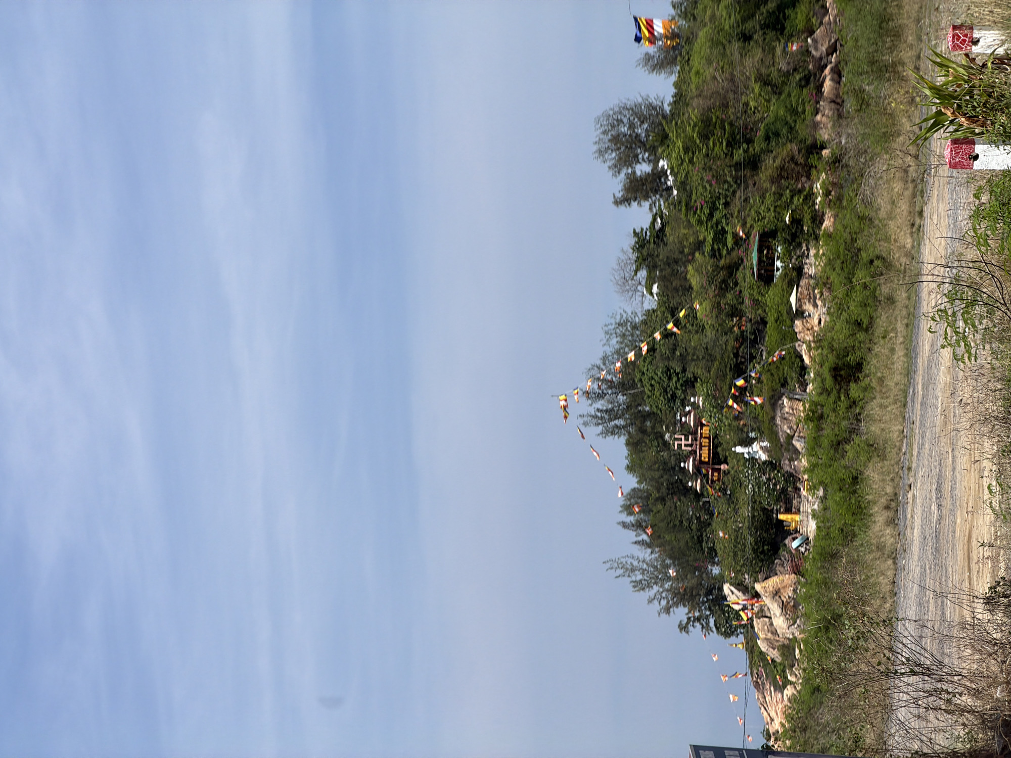 The image depicts a view of a hillside adorned with a Buddhist temple, several flags, and lush green vegetation. The temple structure, visible in the mid-ground, features a prominent swastika symbol on its facade, along with Vietnamese text below it. Prayer flags in various colors, predominantly yellow, red, and white, are strung across the hillside, suggesting a religious or ceremonial context. The hillside itself is rocky and covered with trees and shrubs. In the foreground, a dirt road or path is visible, with some dry grass and a few white and red striped posts. The sky is clear and blue with some wispy clouds. The scene appears to be located in a coastal or elevated area, possibly part of a religious sanctuary. The general ambiance is serene and spiritual, characteristic of a place of worship in Vietnam.