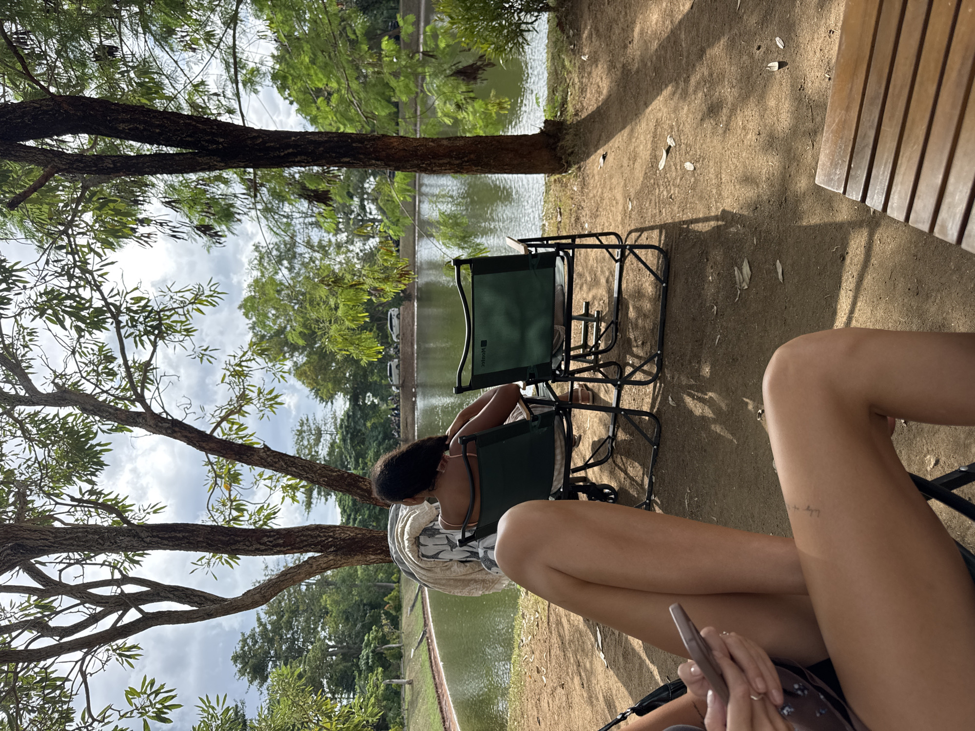 The image depicts an outdoor scene at a park with a body of water. In the foreground, the legs of two individuals are visible, one holding a smartphone. Behind them, a woman sits in a green folding chair, facing the water, with a baby stroller next to her. Another identical green chair is positioned nearby. The background features a tranquil lake surrounded by lush greenery and trees. The sky is cloudy, indicating overcast weather. The location is identified as Kuta Selatan, Indonesia, suggesting a park or recreational area within the region. The overall atmosphere is relaxed and serene, with elements of nature and human presence.