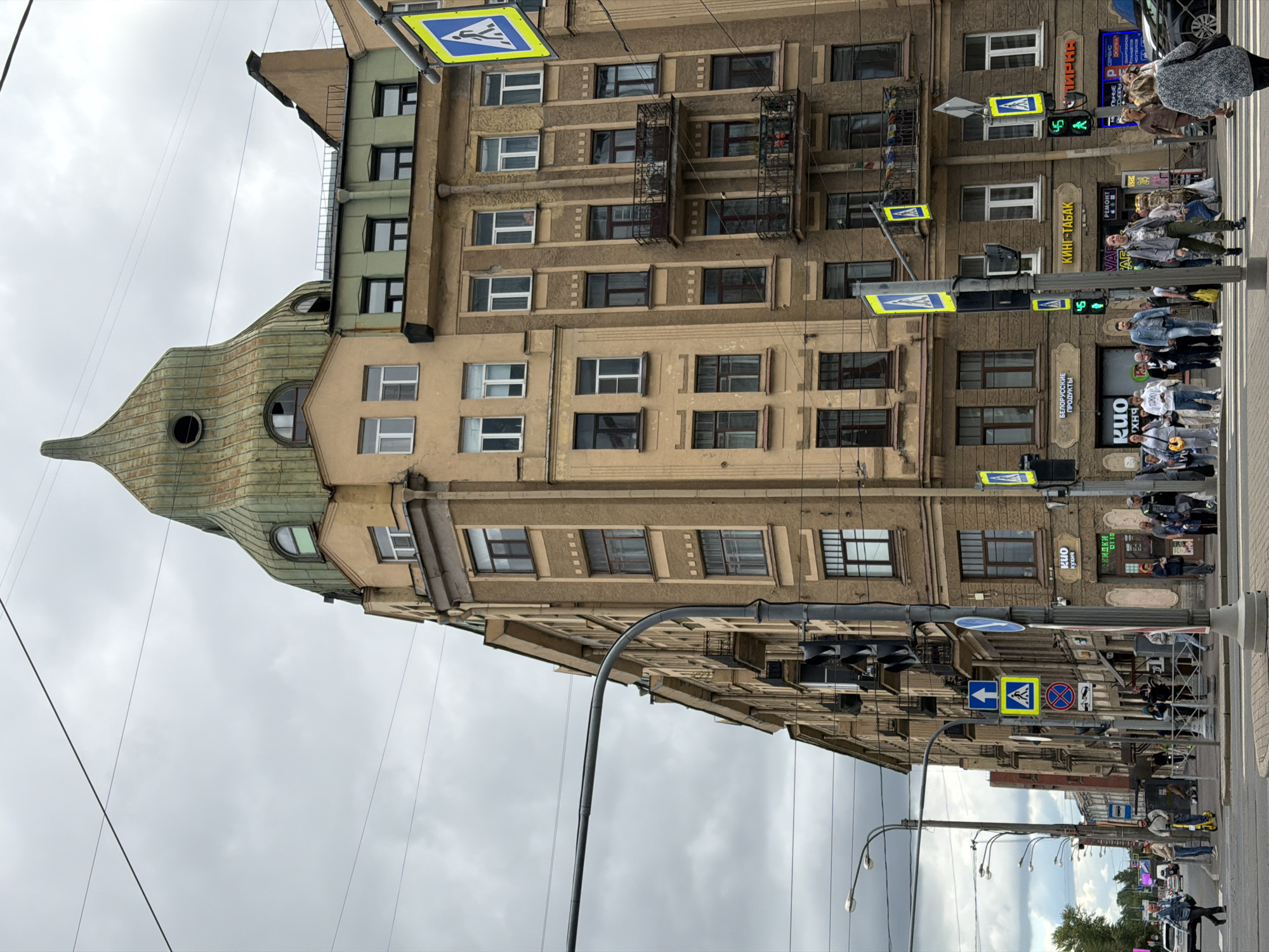 The image captures a street scene in Saint Petersburg, Russia, with a focus on an eclectic architectural ensemble dominated by a tall, tan-colored building featuring a distinctive green, onion-shaped dome with a pointed finial. The building has numerous windows of varying sizes and shapes, some with balconies. Several streetlights, traffic lights, and pedestrian crossing signs are visible, indicating an active intersection. A pedestrian crossing is present in the foreground. Various people are crossing the street, walking on the sidewalk, and standing near the building. Some shops with signage are visible on the ground floor of the building. The sky is overcast, suggesting a cloudy day. The overall impression is of a busy urban environment with a mix of historical and modern elements.