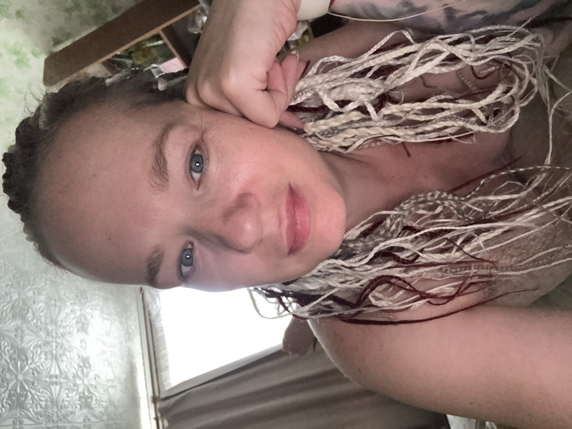 The image shows a close-up, selfie-style portrait of a fair-skinned person with blue eyes and blonde braids, some with red streaks. The person's left hand rests on their cheek. They are partially undressed, with their shoulder visible. The background is blurred and appears to be an indoor setting with patterned wallpaper and shelving. The lighting suggests it is daytime. There is no discernible indication of a location in Moscow, Russia.