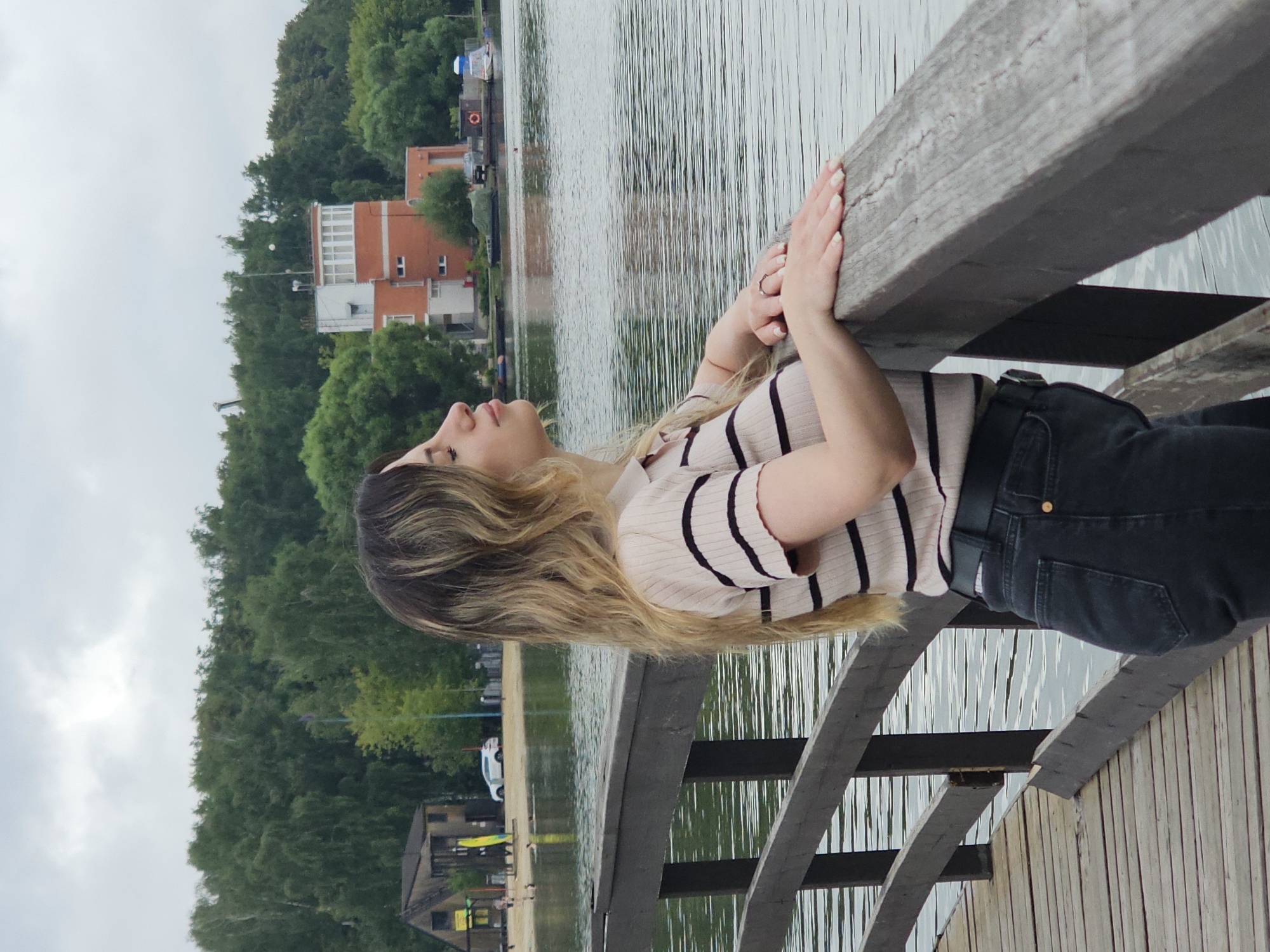 A young woman with long, wavy blonde hair stands on a wooden pier overlooking a body of water. She is wearing a light-colored, striped polo shirt and dark jeans. Her eyes are closed and her head is tilted back slightly, suggesting she is enjoying the moment. Her hands rest on the wooden railing of the pier. In the background, a tree-lined shore is visible, with a few buildings, including a red and white structure that appears to be a watchtower or control tower, and a smaller wooden building. Boats are scattered on the water, and there is a sandy area with people near the shore in the distance. The sky is overcast. The scene is likely a recreational area within Moscow, Russia.