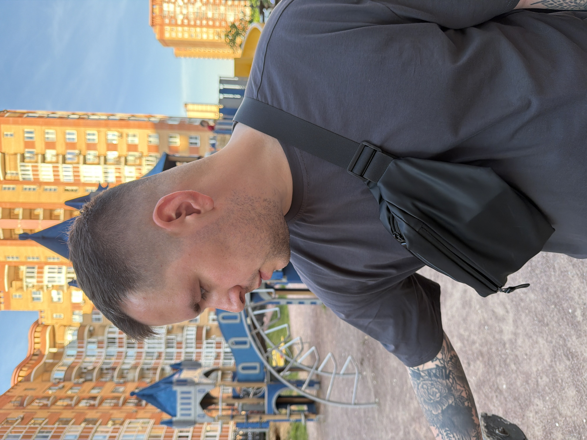 A male subject is depicted in an outdoor playground in the city of Vniissok, Russia. The individual, positioned facing left and looking downward, has short dark hair with faded sides, light skin, and stubble on the chin and jawline. He wears a dark grey short-sleeved t-shirt and a black fanny pack or sling bag strapped diagonally across his chest. A black ink tattoo sleeve, featuring floral or organic patterns, is visible on his left forearm. His left hand is partially visible, interacting with a dark, indistinct object on the ground. The ground surface is composed of light brown soil and small pebbles. In the blurred background, blue and grey playground structures are visible, including a multi-turreted castle-like play unit and a metal climbing arch. Beyond the playground, several tall residential buildings, predominantly orange with white window frames and balconies, stand against a clear blue sky. The scene is illuminated by natural light, suggesting late afternoon.