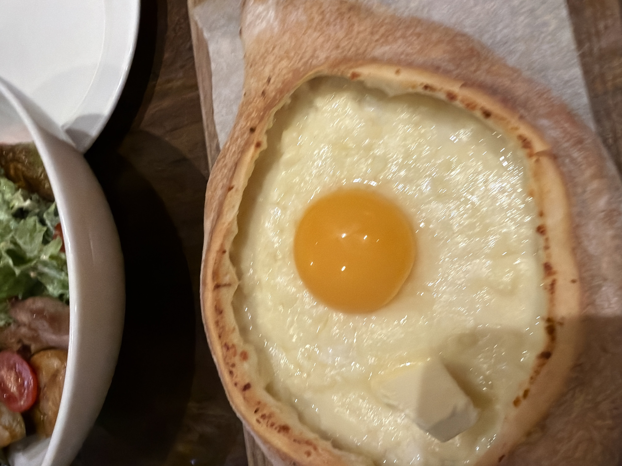 The image shows a partially obscured view of a meal on a dark wooden table. On the left is a white bowl containing a salad with lettuce, sliced tomatoes, and unidentified protein. Above the bowl is a white plate. To the right of the salad is a khachapuri, a Georgian cheese bread filled with a mixture of cheese, an egg yolk, and a pat of butter. The khachapuri sits on a piece of parchment paper. There are no people visible in the image. There is no specific information in the image that supports a location in the city of Saint Petersburg, Russia.