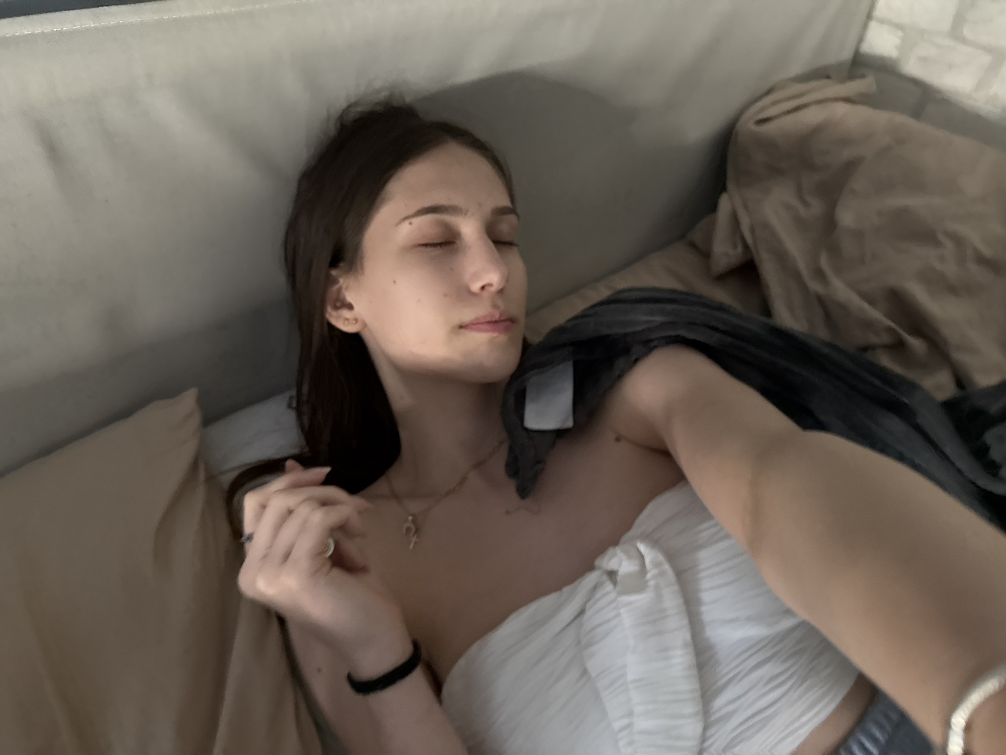 A young woman with long dark hair is lying on a sofa or bed, eyes closed. She is partially covered with a white, textured top or fabric and a dark gray blanket. The sofa is light-colored, and there is a tan-colored blanket or sheet nearby. She is wearing a black wristband on her left wrist and a necklace. She also appears to be wearing earings and rings.
There is no information in this image to determine if this is in Sofia, Bulgaria.